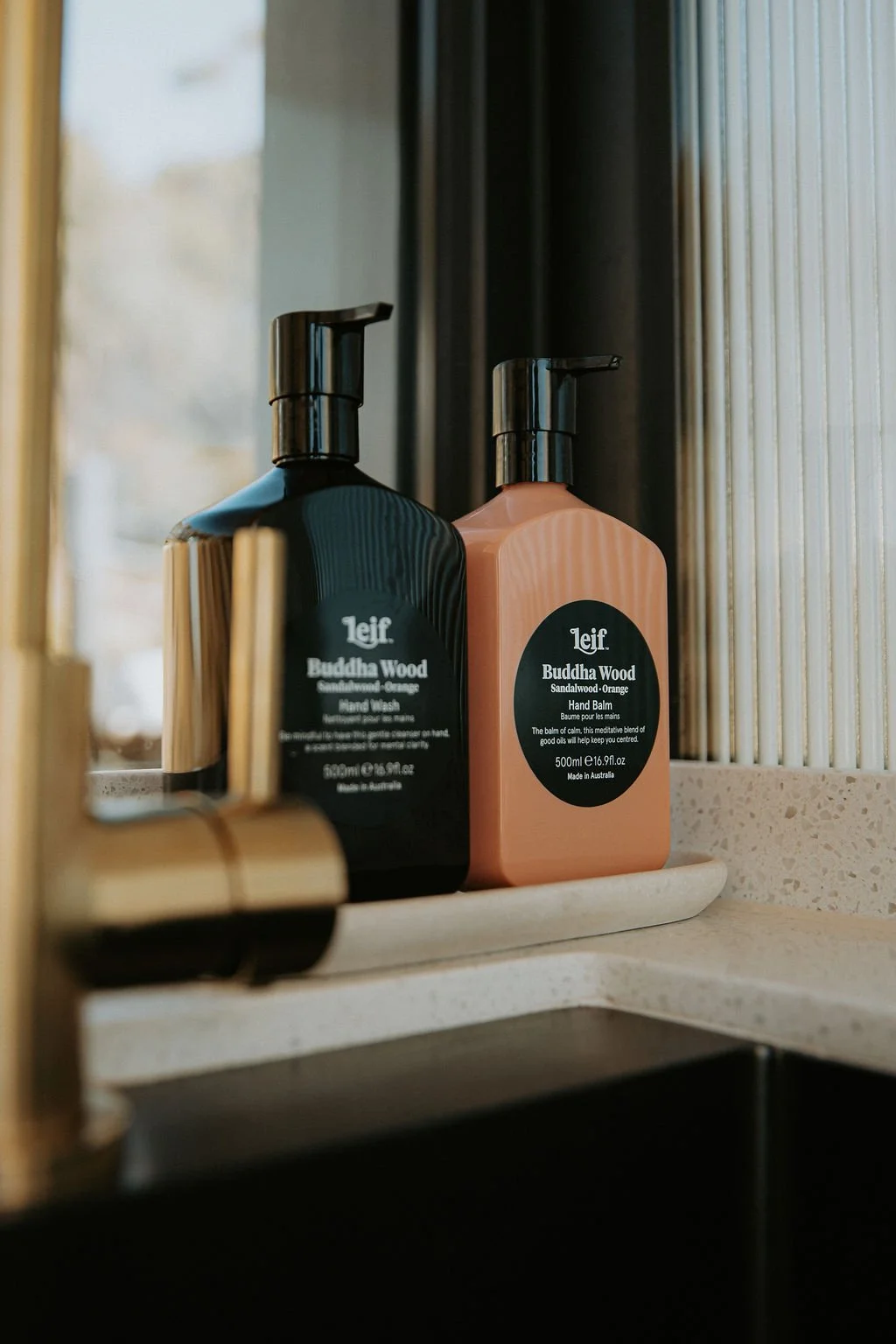 Two bottles of Leif Buddha Wood hand wash and hand balm on a bathroom shelf next to a mirror.