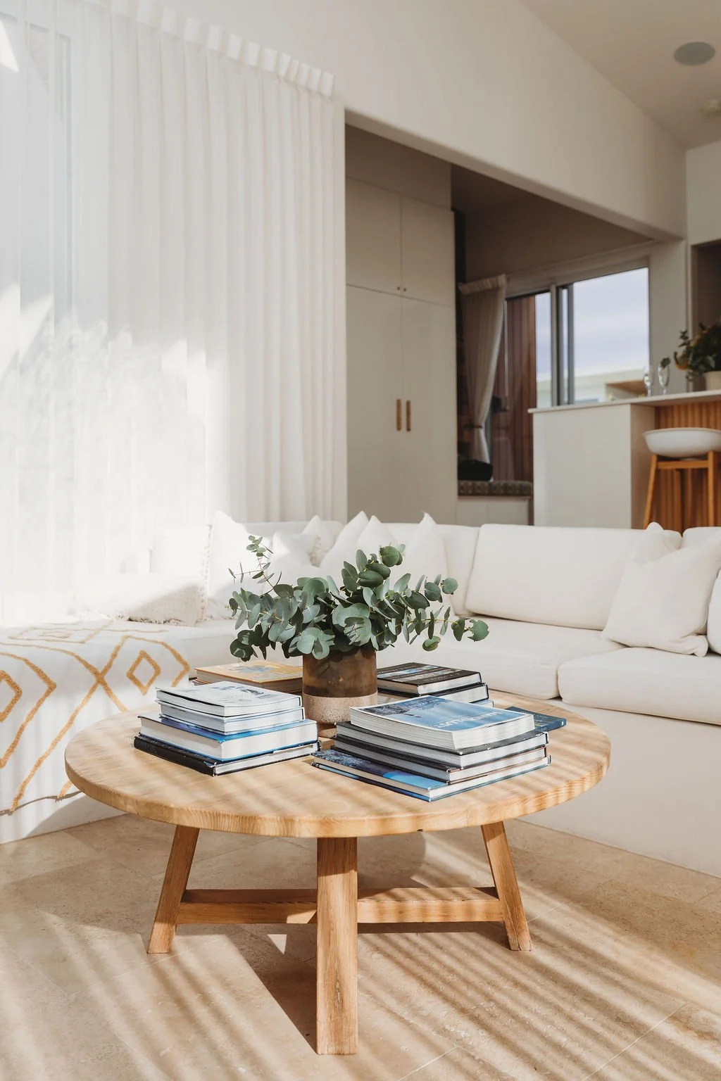 Living room with a round wooden coffee table, stacks of magazines, a vase with eucalyptus, white sofa, and curtains in the background.