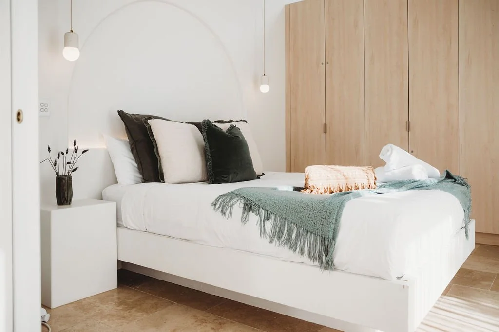 Minimalist bedroom with a white bed, dark and light pillows, green throw blanket, wooden wardrobe, and wall-mounted lamps.