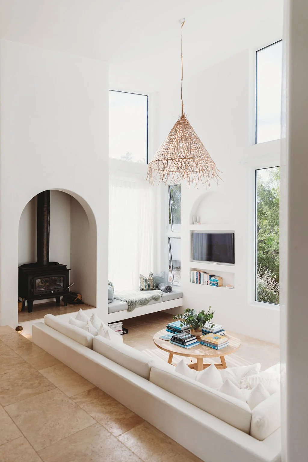 Bright living room with large windows, white walls, a woven pendant light, a black stove in an arched alcove, a built-in bench with pillows, a flat-screen TV, a bookshelf, and a round wooden coffee table with books and a plant.