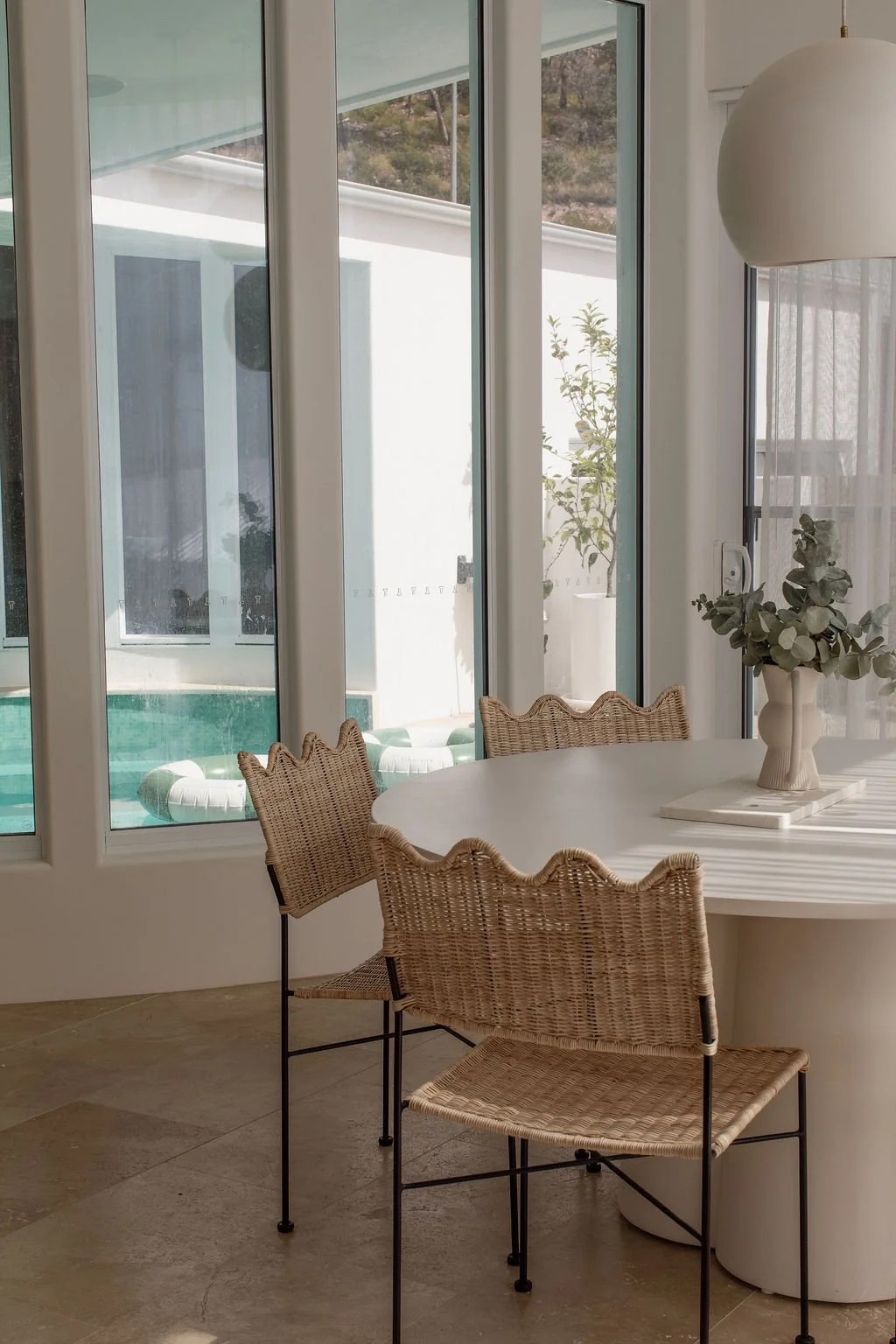 A cozy indoor dining area with a white oval table, wicker chairs, a vase with greenery, and large glass windows showing a pool and outdoor patio with potted plants.
