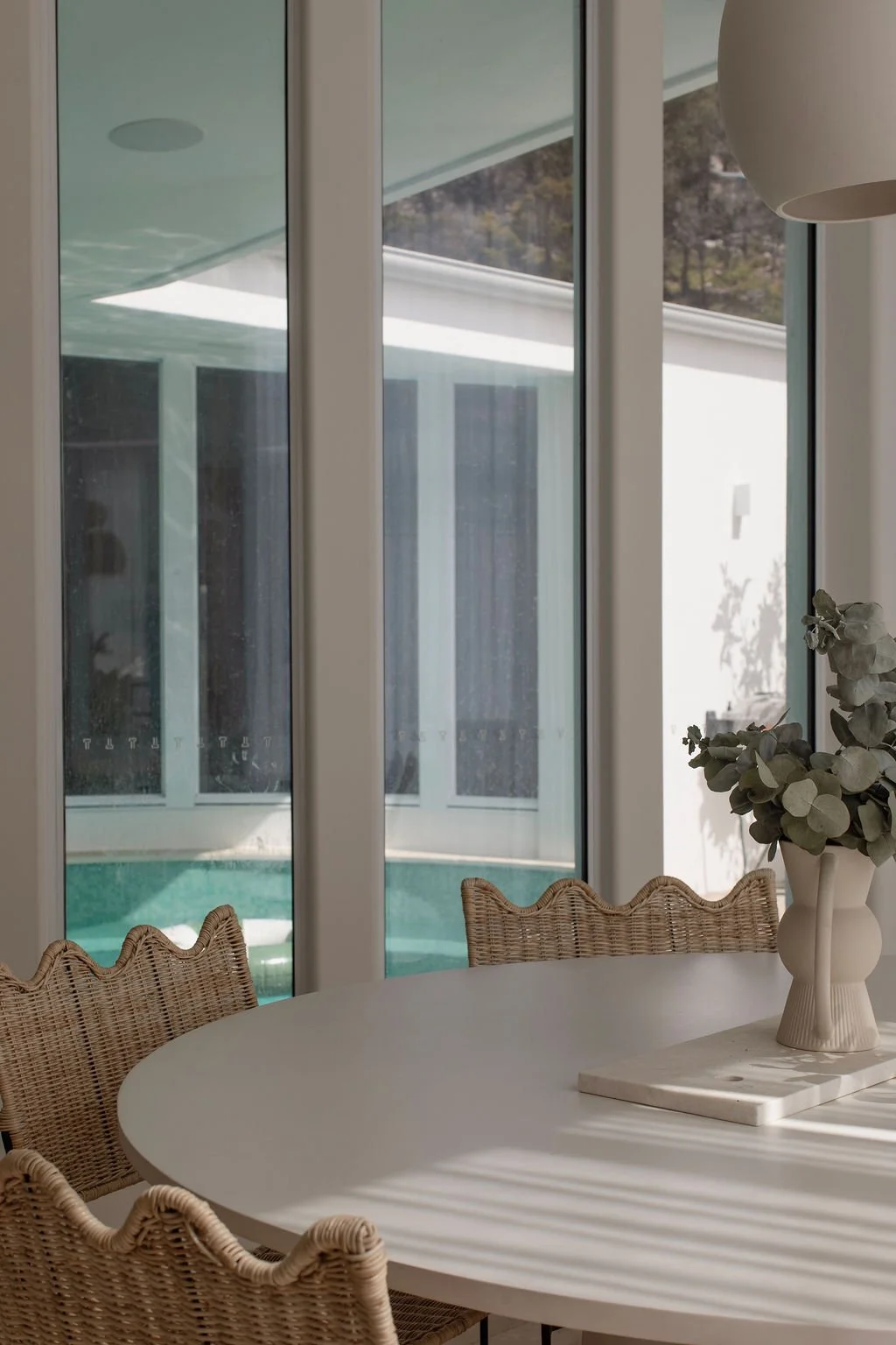 Sunlit dining area with a round white table, wicker chairs, a white vase with greenery, and large glass windows showing a backyard with a pool and white exterior walls.