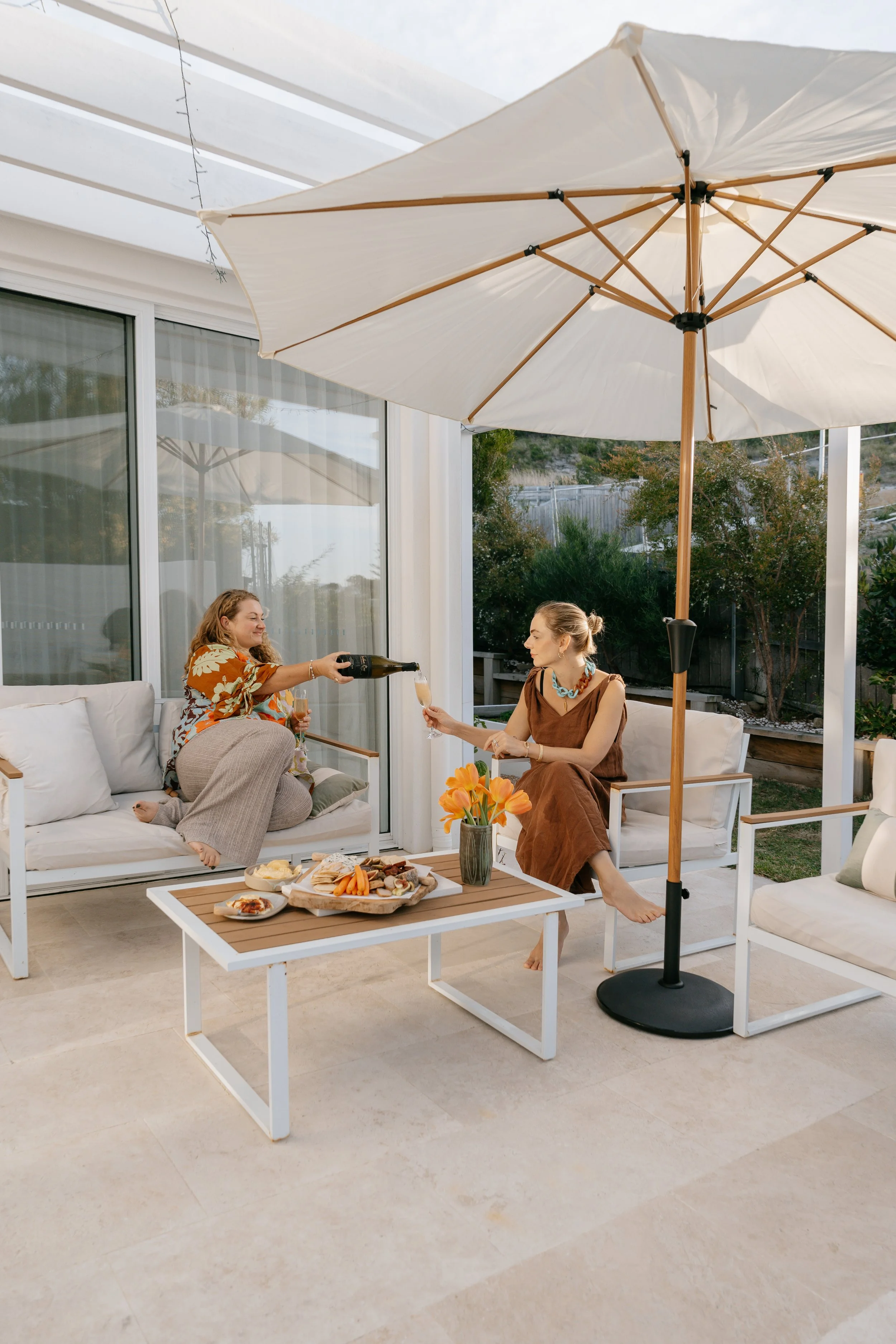 Two women enjoying a sunny outdoor gathering under a large umbrella, with a table of snacks and drinks nearby.