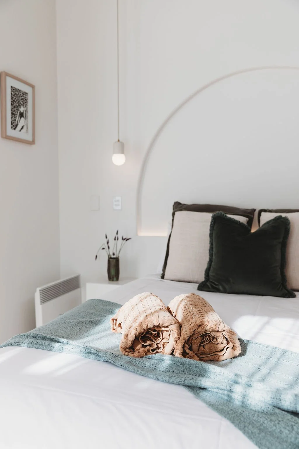 A neatly made bed with rolled beige towels on top, in a minimalistic bedroom with neutral-colored pillows, wall art, a pendant light, and a small vase with dried flowers on a nightstand.
