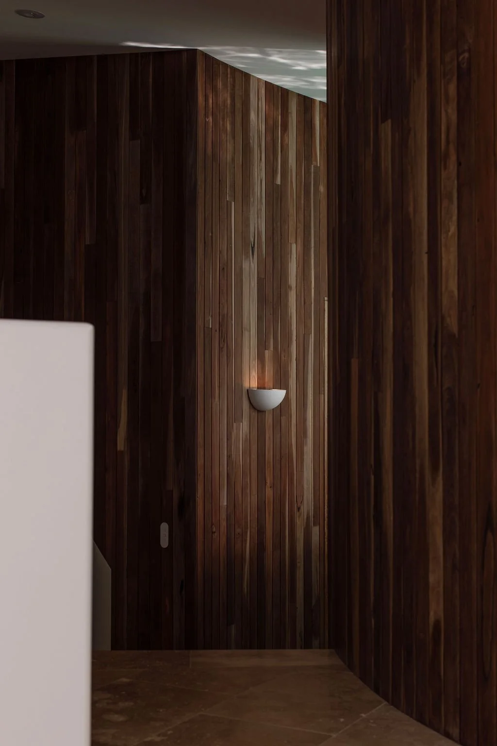 Interior view of a room with wooden paneling on the walls, a white wall sconce light fixture, and part of a white appliance or furniture in the foreground.