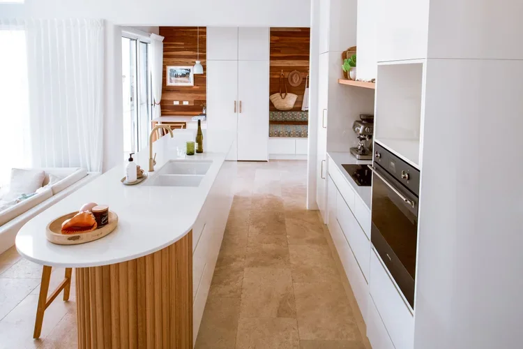 Modern kitchen with white cabinets, a island with a wooden base, and built-in oven. There are large windows letting in natural light, with a cozy sitting area visible in the background.