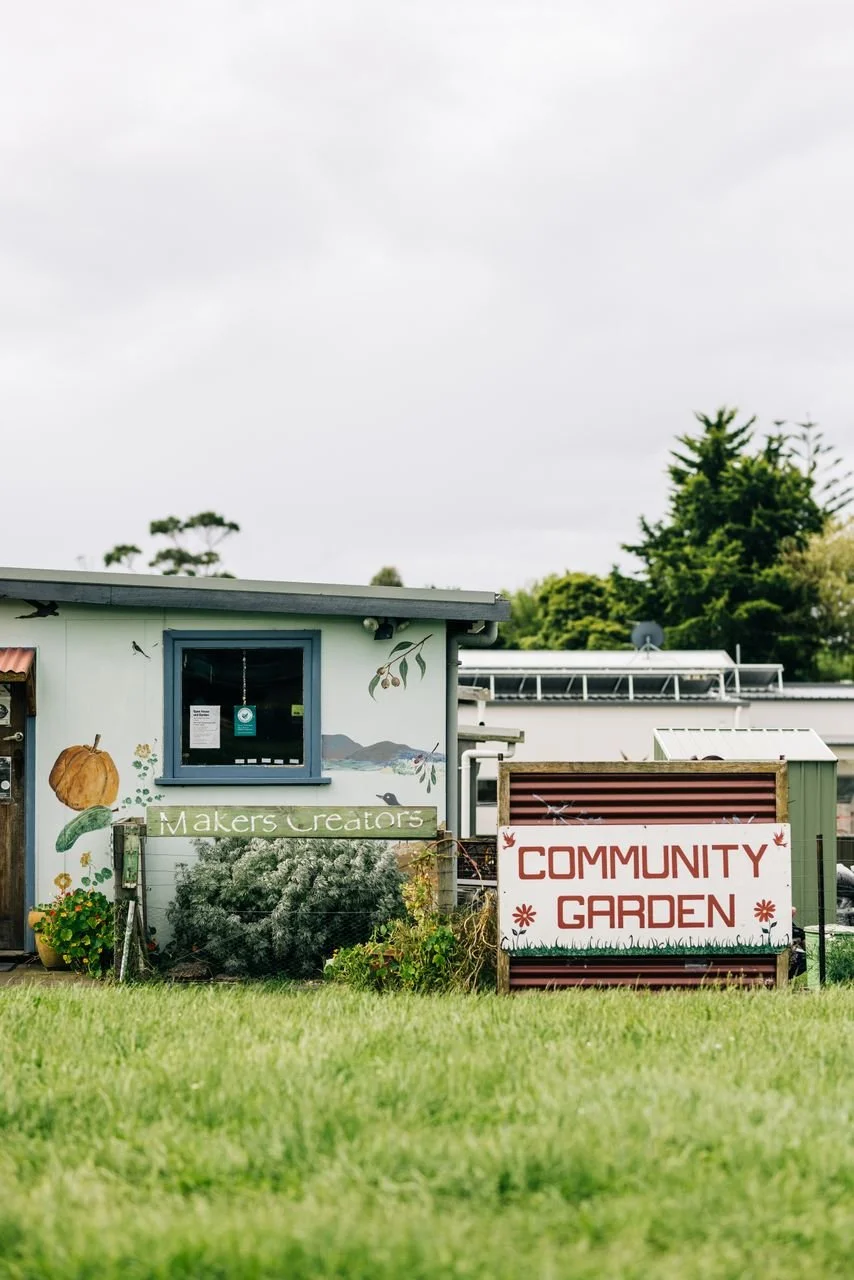 Community garden with a sign reading 'Community Garden' and a building with a mural, surrounded by grass and trees.