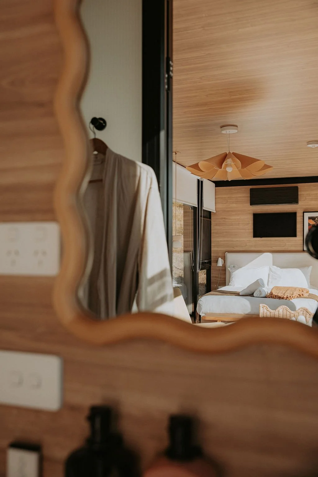 A bedroom with wooden walls and ceiling, featuring a bed with white linens, towels, and pillows, a wall-mounted TV, a side table with a lamp, a modern ceiling light fixture, and a sliding glass door leading outside. The photo is taken through a mirro