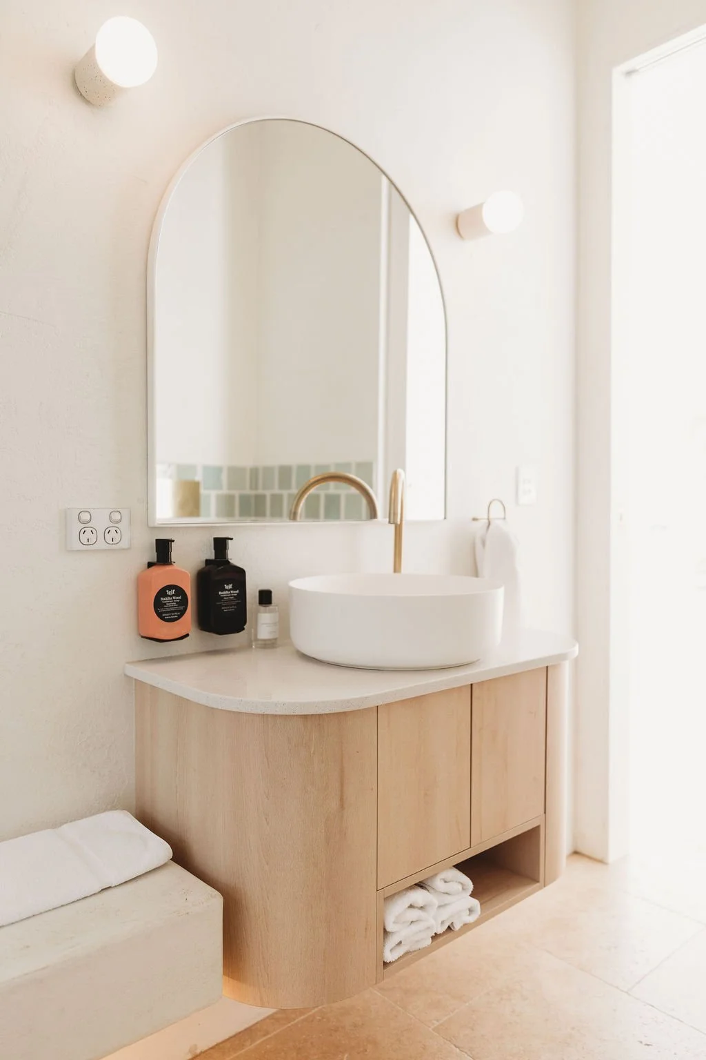 Modern bathroom vanity with a round vessel sink, gold faucet, large wall mirror, two wall-mounted lights, and two soap bottles on the countertop. Wooden cabinet with open shelf containing folded towels. Wall outlet and a towel ring with a towel.
