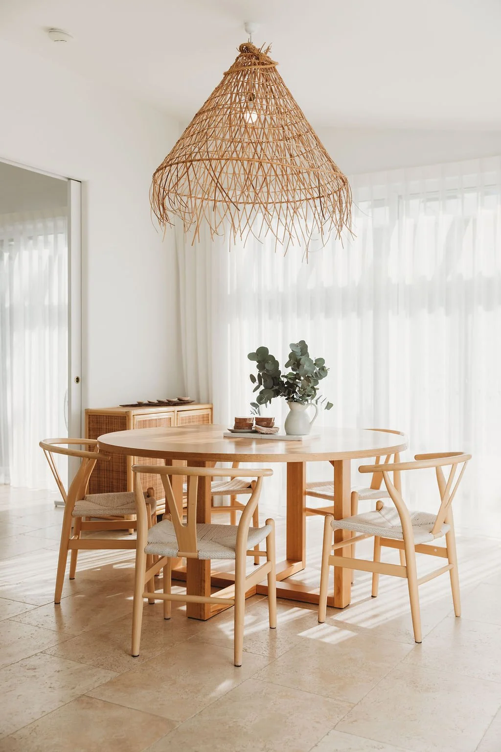 Bright dining room with a round wooden table, four matching wooden chairs with woven seats, a white vase with green eucalyptus, and a large woven pendant light hanging from the ceiling.