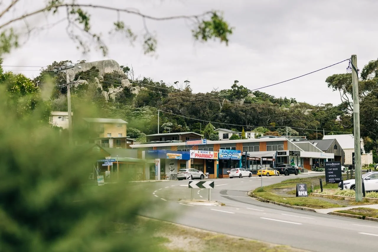 A small town street with shops including a pharmacy, a bakery, and a restaurant, with cars parked along the road and a large hill or mountain in the background under a cloudy sky.