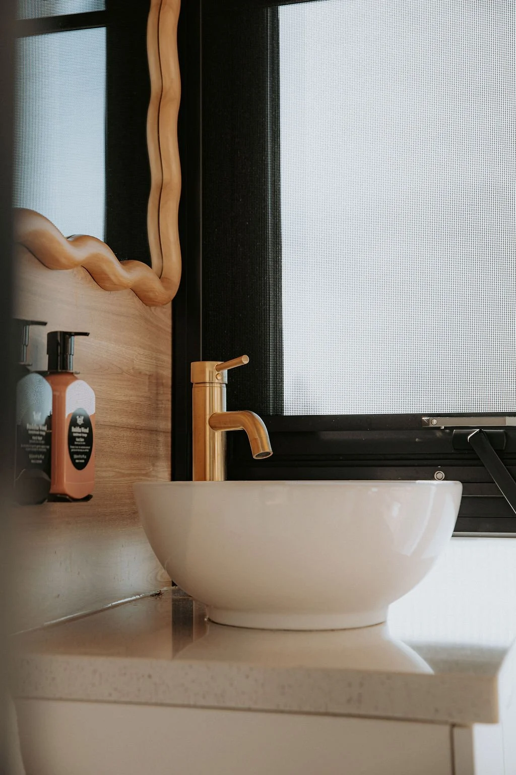 Bathroom sink with gold faucet, mirror with wavy wooden frame, soap bottles, and window in the background.