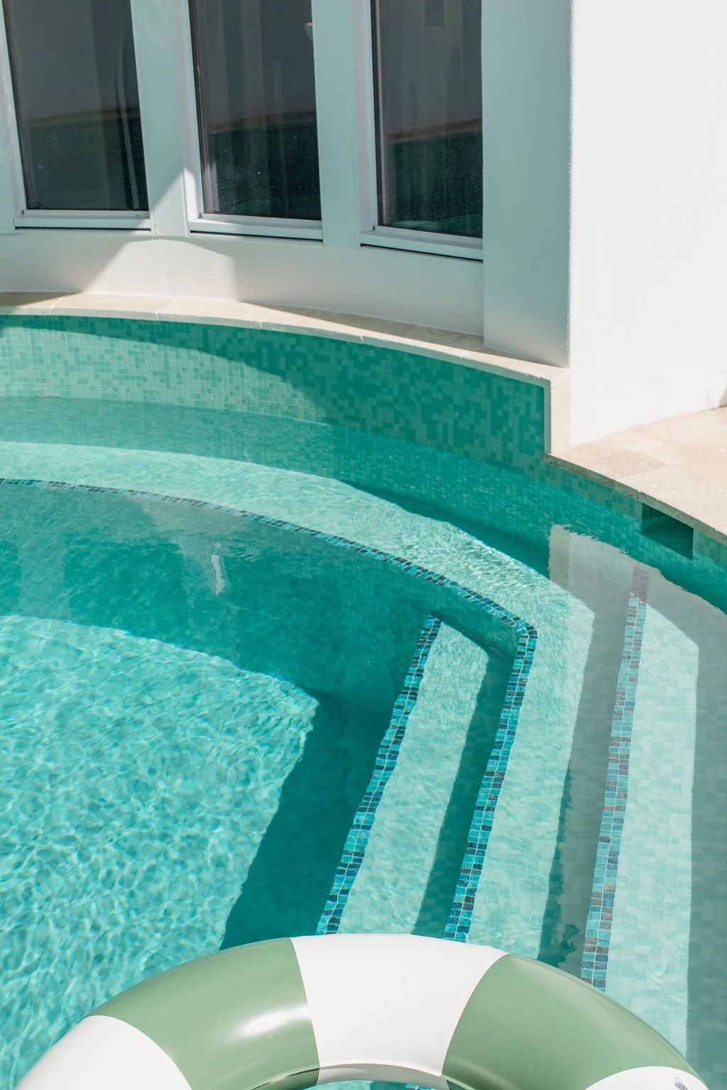 Part of a swimming pool with built-in steps, tile border, and a floating life preserver near the edge, adjacent to a building with large windows.