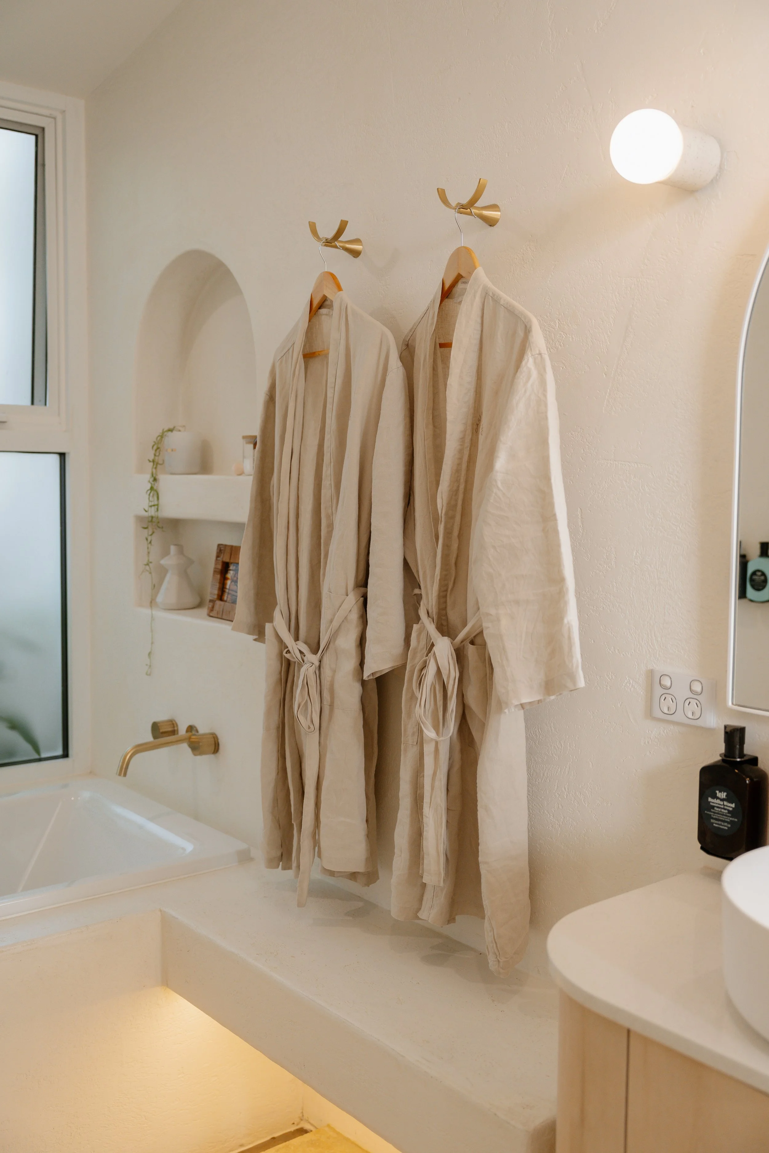 Living space with two beige robes hanging on wall hooks, a small white shelf with decorative items, a window, and a sink in the foreground. The room has a soft, neutral color scheme with warm lighting.