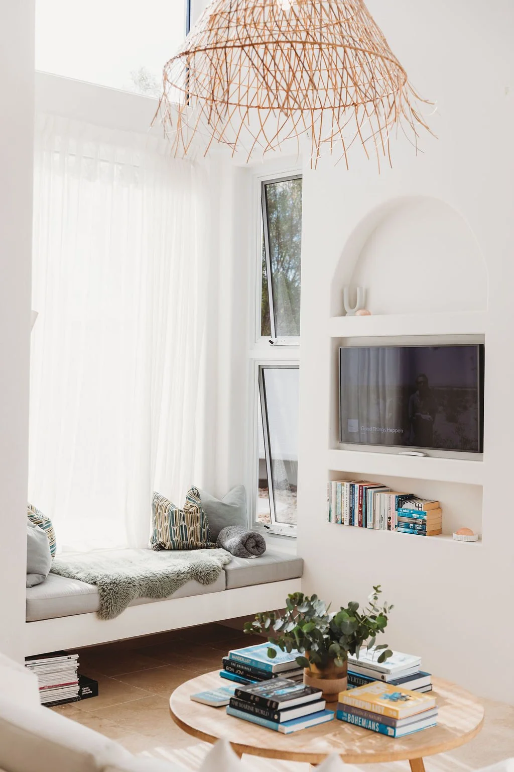 Cozy living room with white walls, a large window with sheer curtains, a built-in window seat with pillows and a throw blanket, a wall-mounted TV, a bookshelf below the TV, and a wooden coffee table with books and a plant.