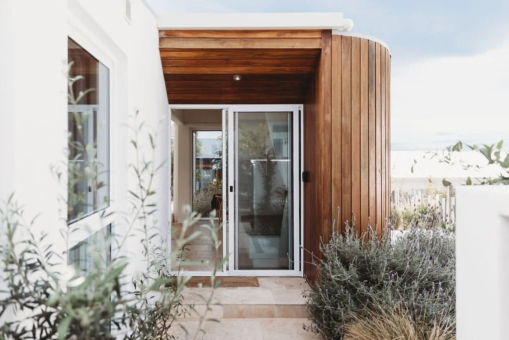 Modern house entrance with sliding glass door, wooden curved wall, and a front porch surrounded by desert plants.