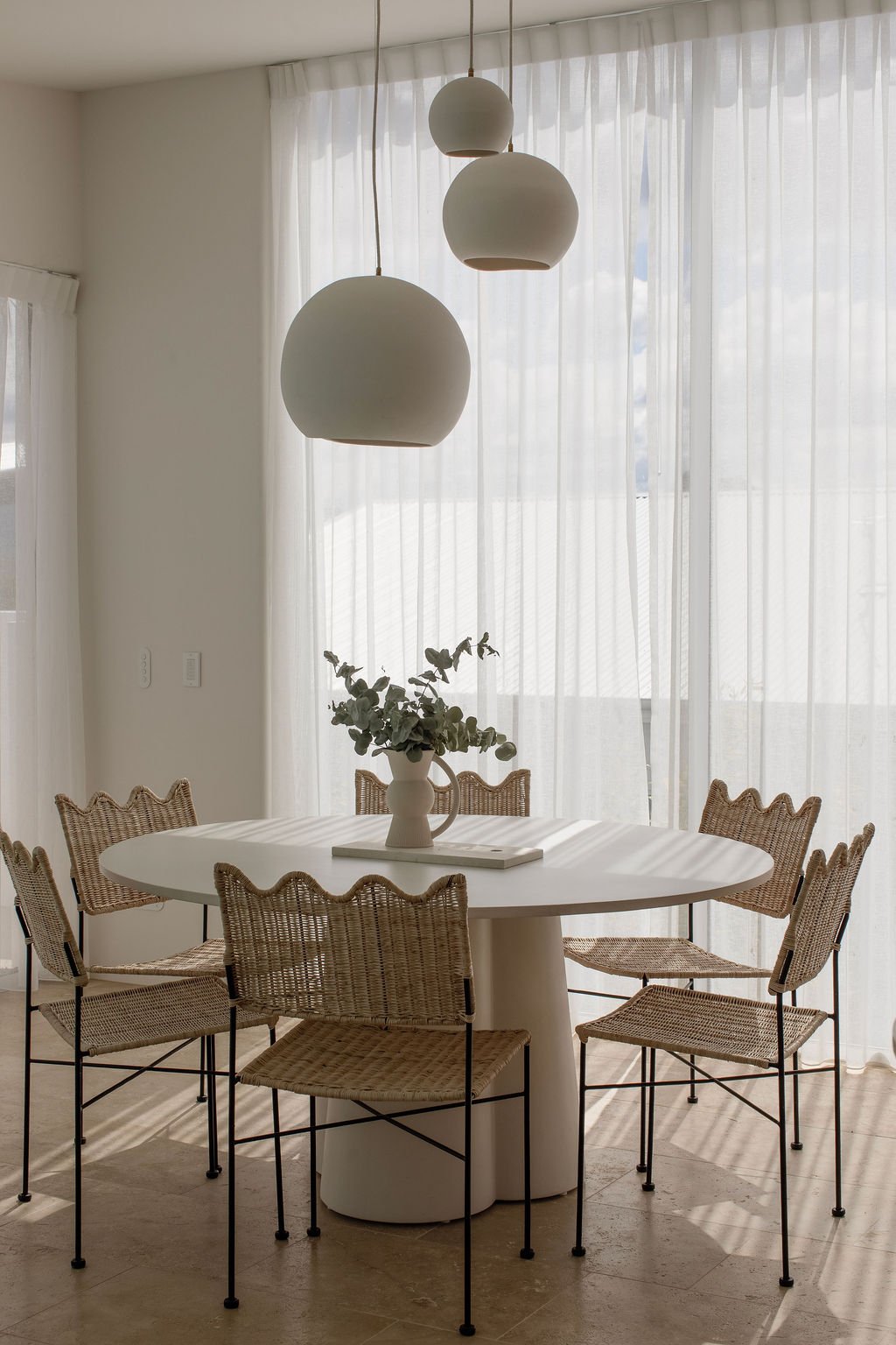 A modern dining room with a white oval table surrounded by six wicker chairs with black metal legs. A white vase with eucalyptus leaves is on the table. Three large white pendant lights hang from the ceiling. The room has sheer white curtains letting