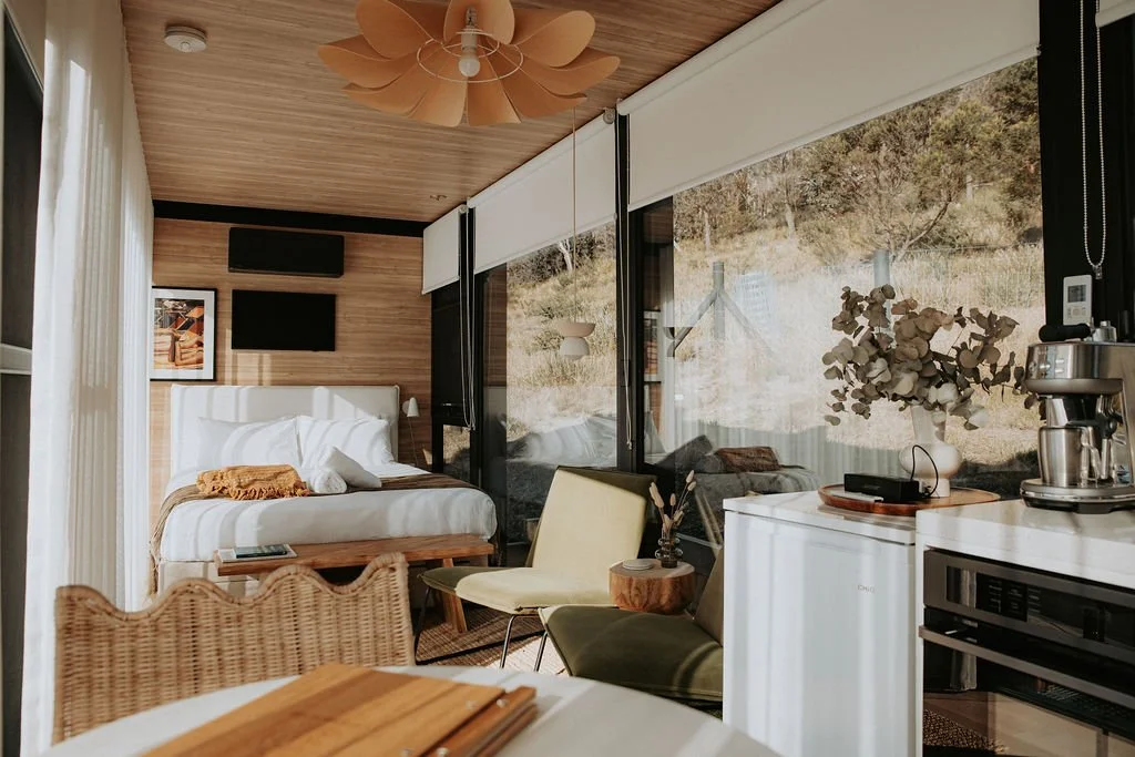 A modern bedroom with a large bed, wooden headboard, and white bedding. A small wooden table and a wicker chair are nearby. There are large windows with views of trees and outdoors. A ceiling fan, a framed picture on the wall, a TV, and a kitchenette