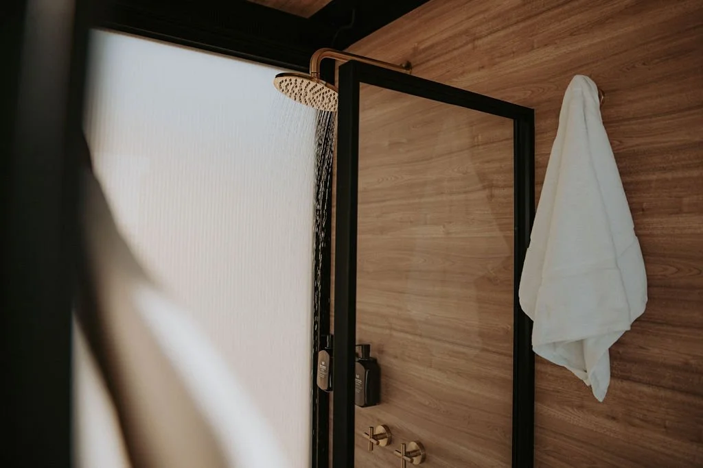 Shower area with a black-framed glass door, a copper showerhead on a wooden wall, and a white towel hanging on a hook.