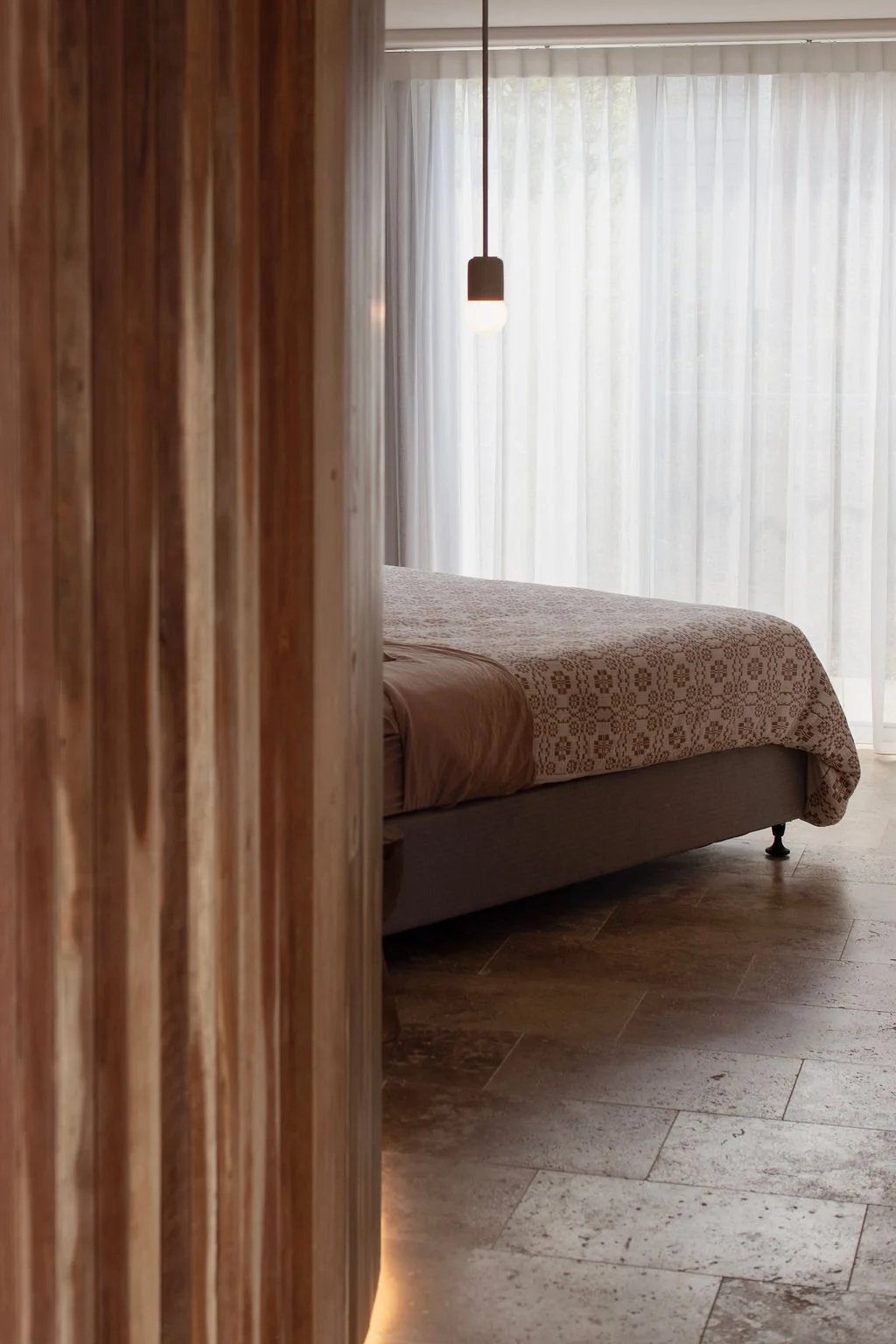 A bedroom with a bed that has a patterned quilt, a hanging pendant light, and large window with sheer white curtains, seen through a partially open wooden door.