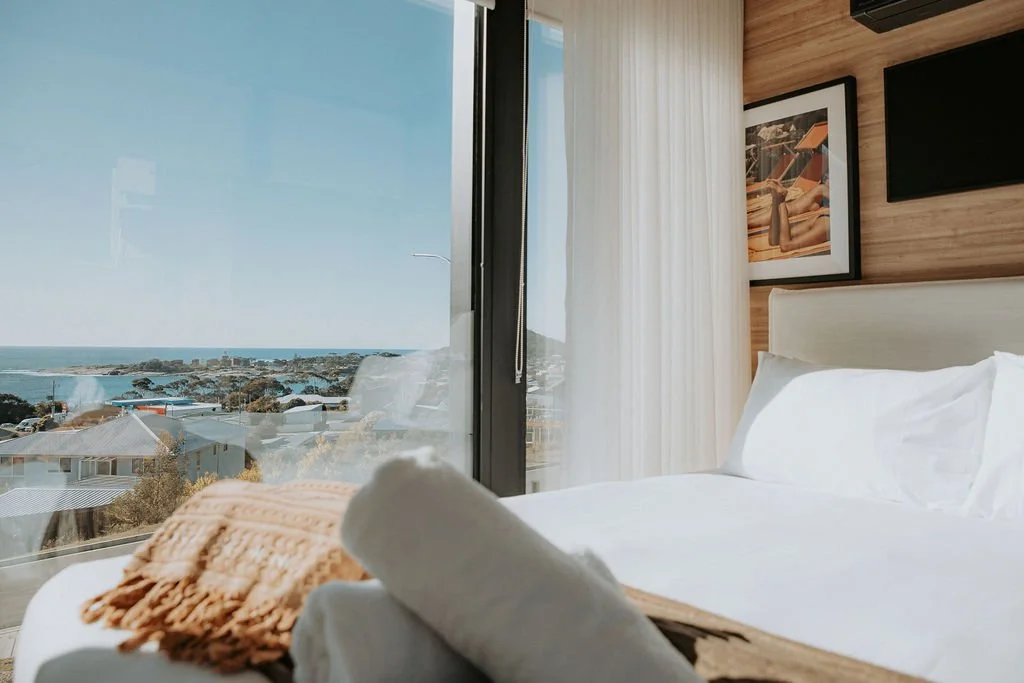 A bedroom with a large window overlooking a scenic coastal landscape, with clouds and the ocean in the background. The room features a white bed with pillows, beige throw blanket, framed art on a wooden wall, and light-colored curtains.