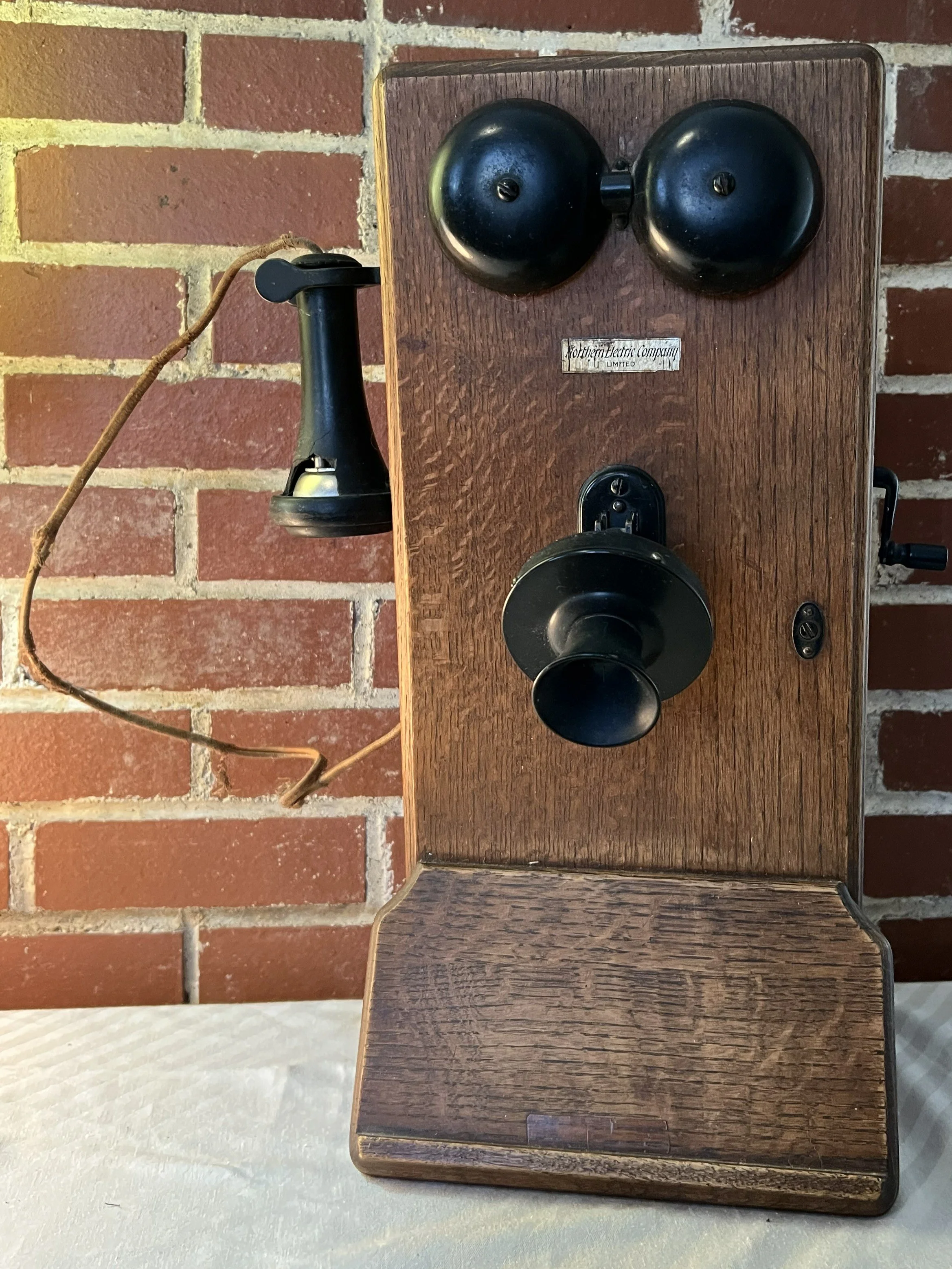 A vintage wall-mounted telephone with a wooden body, black rotary dial, and a separate black receiver hanging on the side, set against a red brick wall.