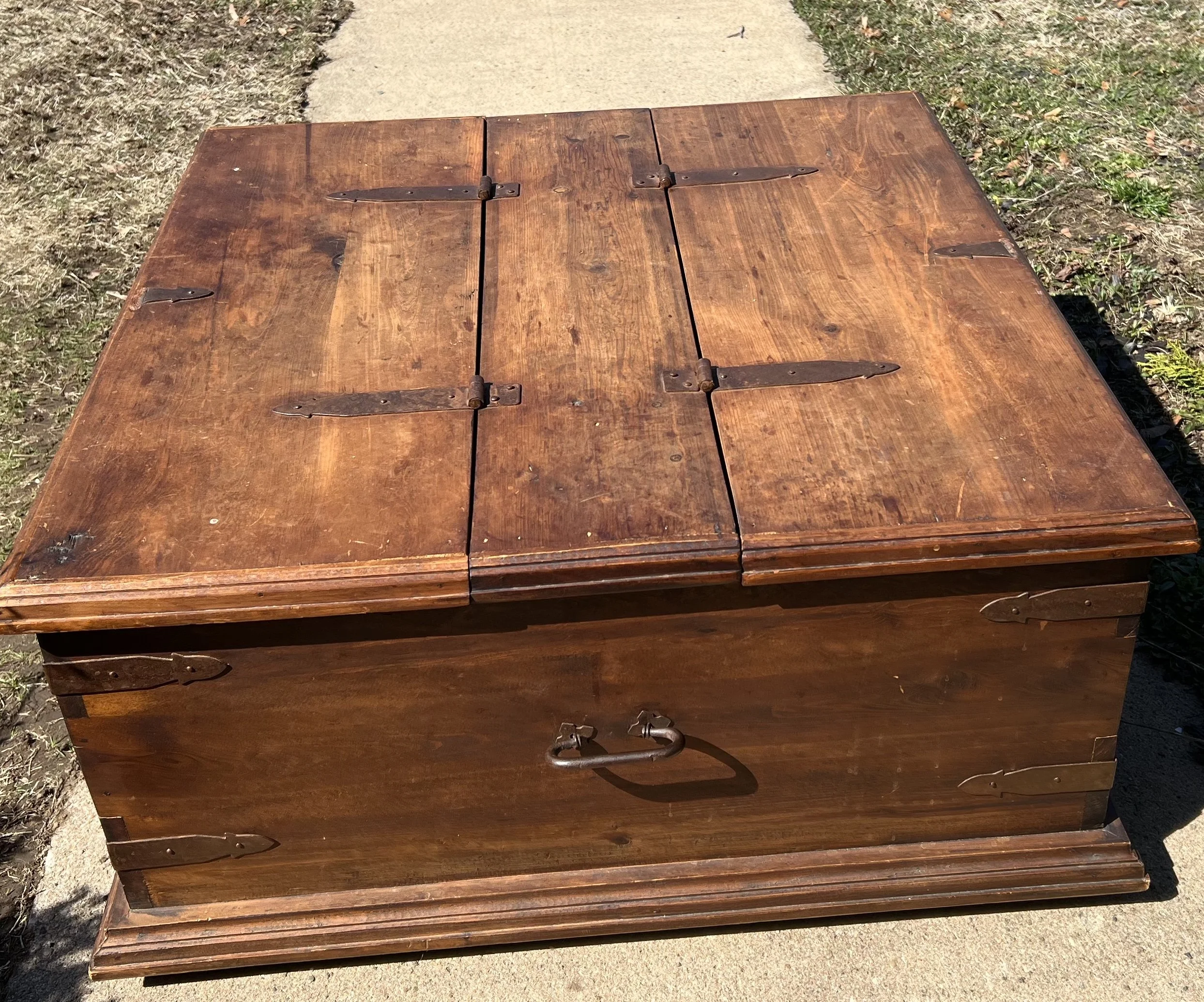 Vintage wooden chest coffee table crafted from solid wood. It features a rustic design with heavy iron hinges and metal handles, common in mid-to-late 19th-century styles. 
A square or rectangular "trunk-style" coffee table with a split-opening lid a