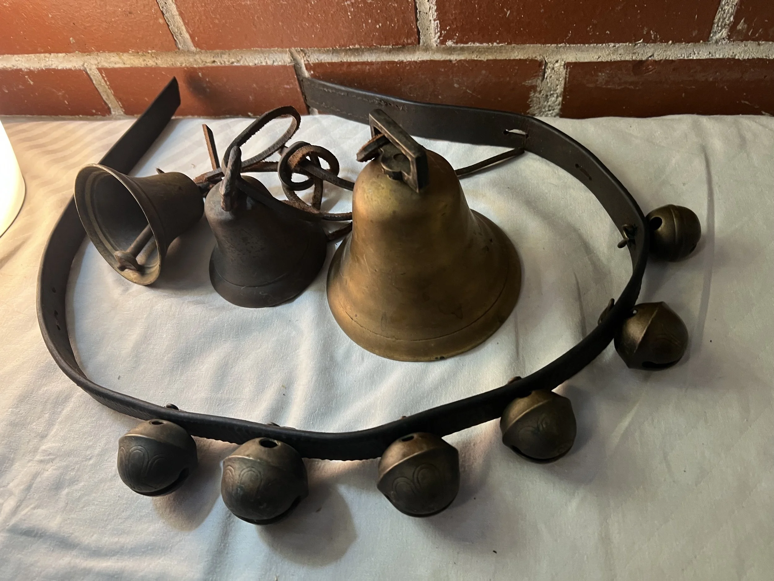 A vintage wall-mounted bell system with a large brass bell, a smaller bell, a frame with multiple small bells, and a wall bracket, on a white cloth surface against a brick wall.