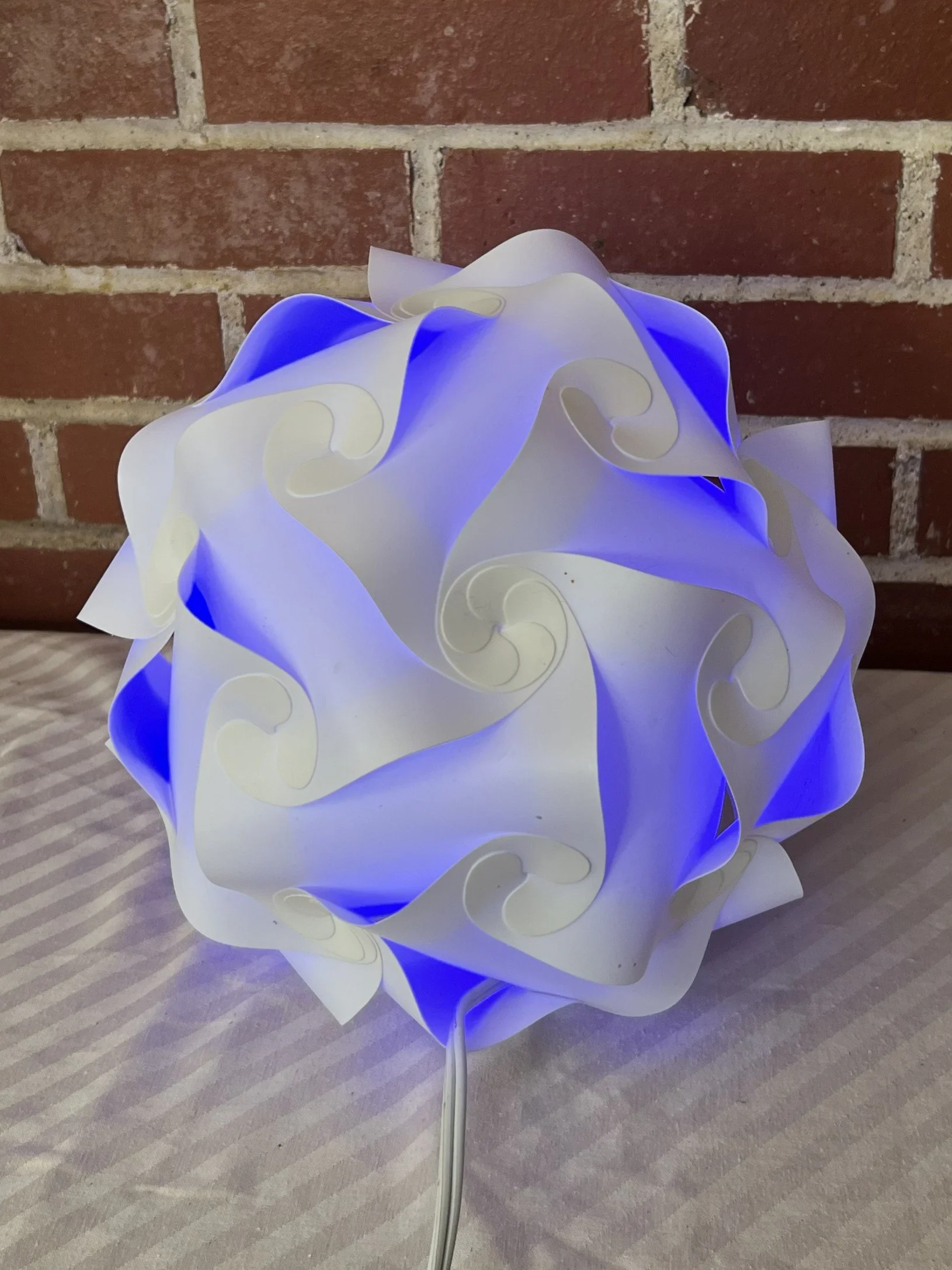 A decorative table lamp with a spherical shape made of white and blue swirling, petal-like plastic panels, illuminated from inside, placed on a table with a brick wall in the background.