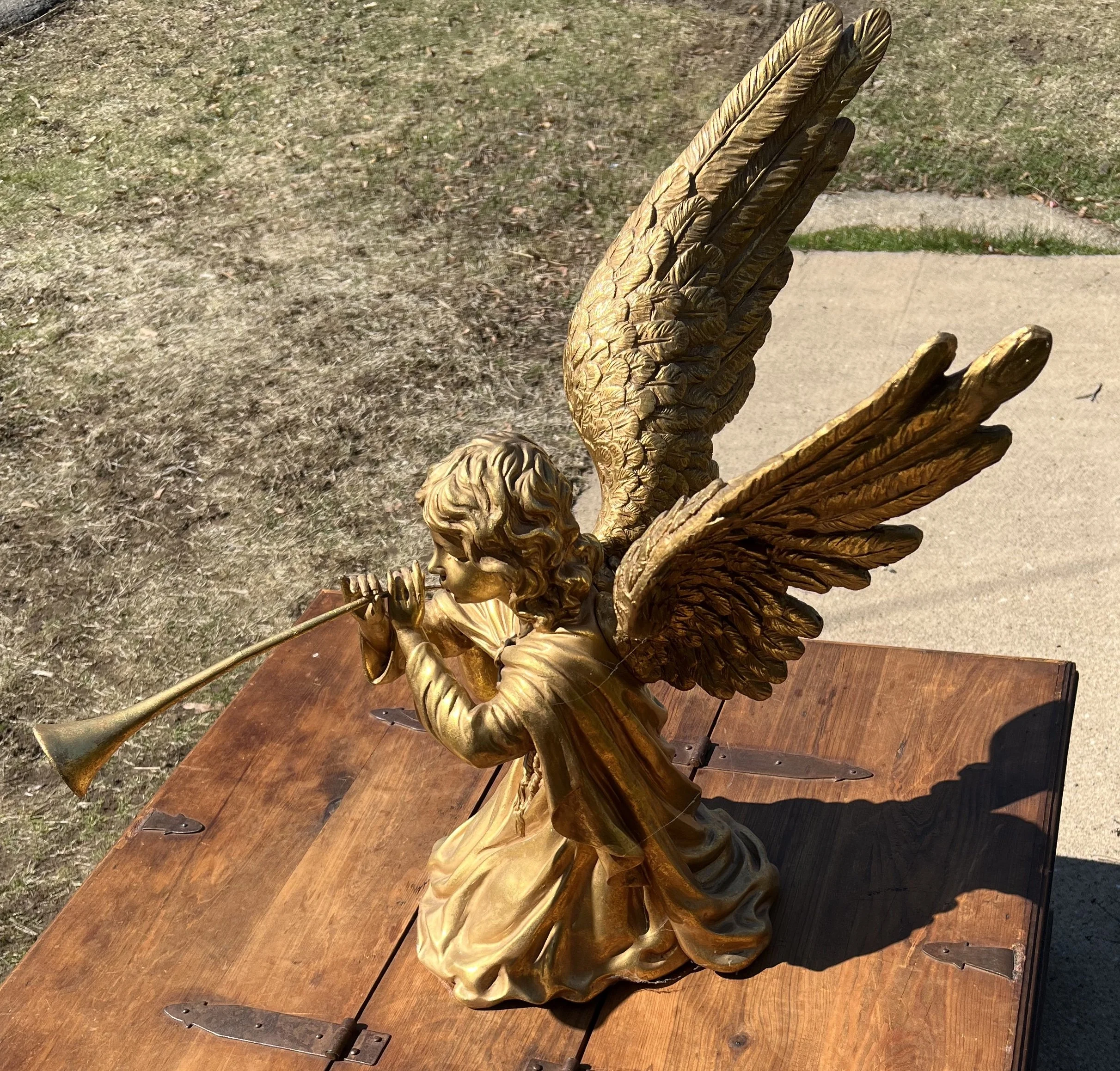 Polystone angel playing a trumpet. 
 It is constructed from sturdy poly-resin designed to look like bronze.
The figure stands 24 inches tall. The angel is hand-painted in a gold finish and features detailed, spread wings.