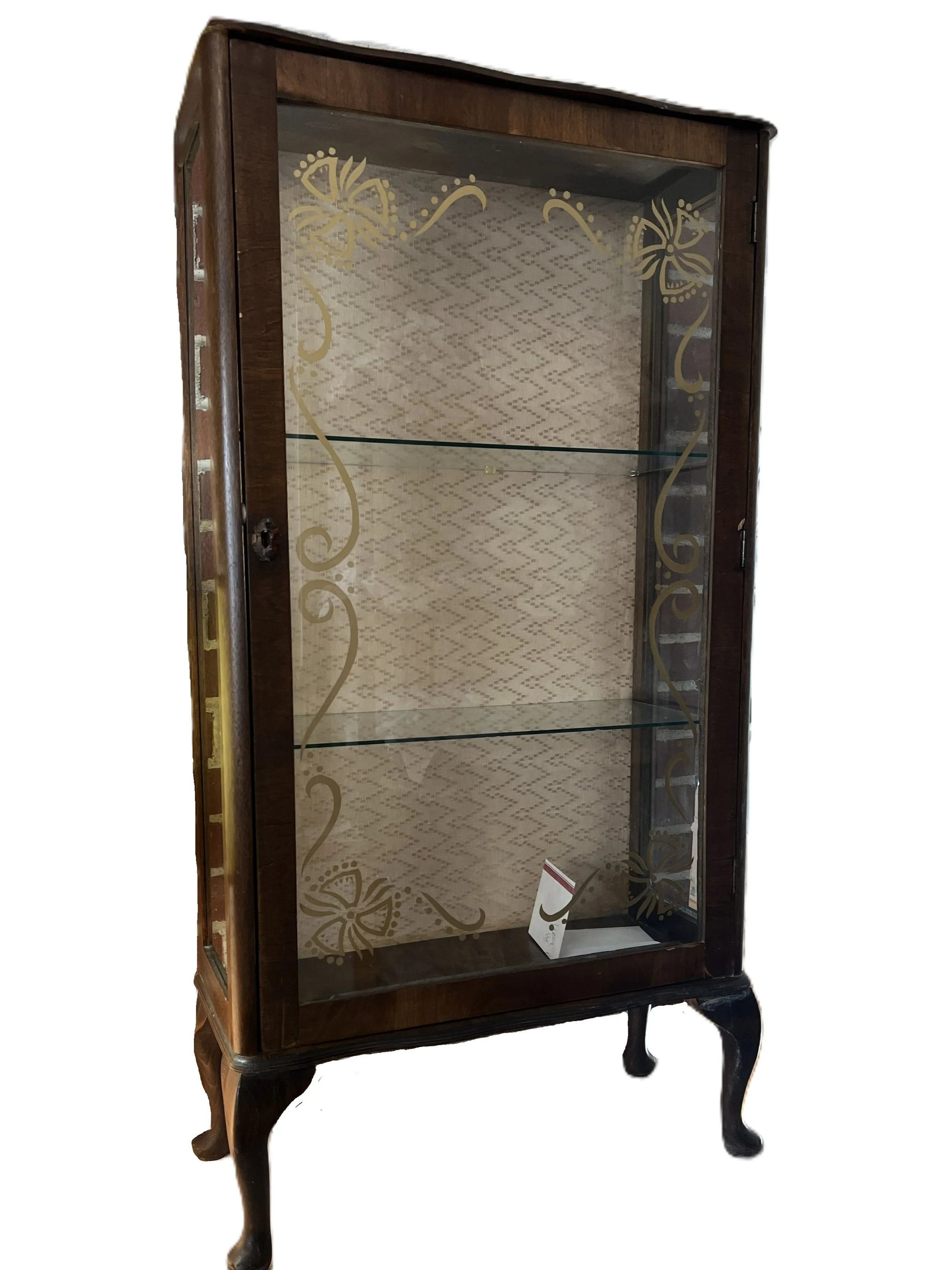 A vintage wooden display cabinet with glass doors decorated with gold floral patterns, standing on four curved legs with a patterned interior backdrop and three glass shelves.