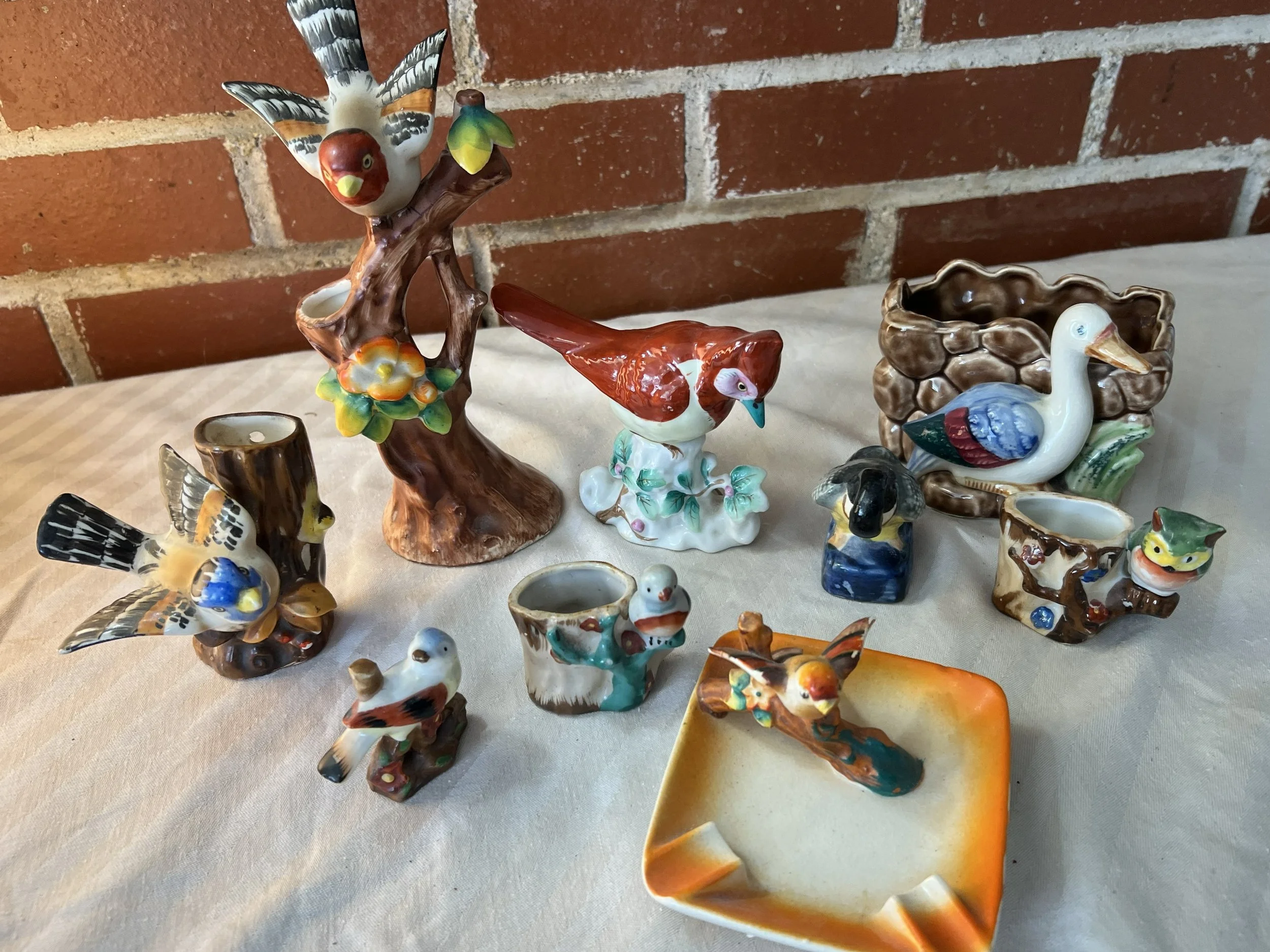 Collection of ceramic animal figurines, including birds, a duck, and a frog, arranged on a table with a brick wall background.