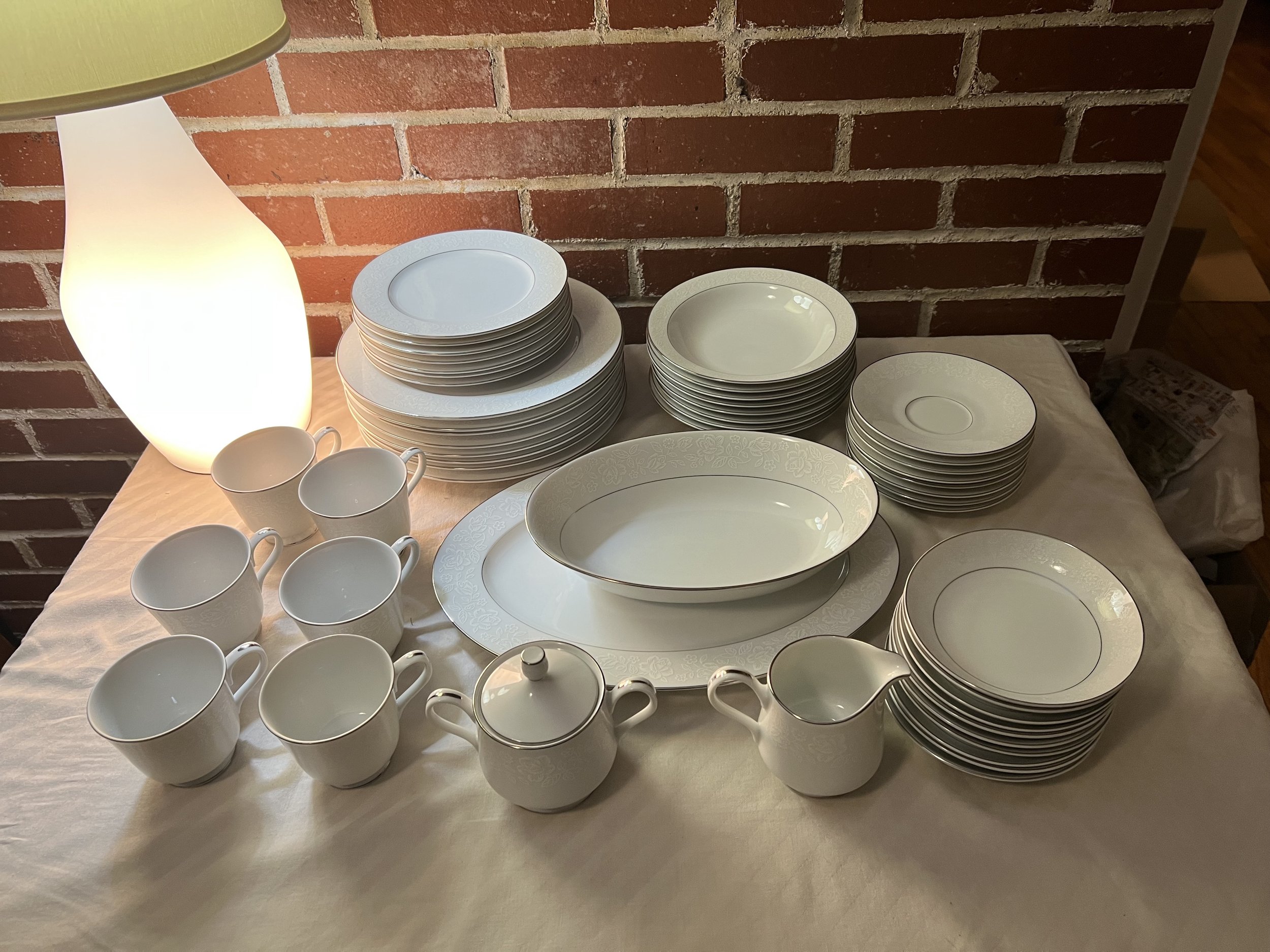A table with a set of white ceramic dinnerware including dinner plates, salad plates, bowls, mugs, a teapot, a creamer, and a large oval serving dish, illuminated by a table lamp against a brick wall background.