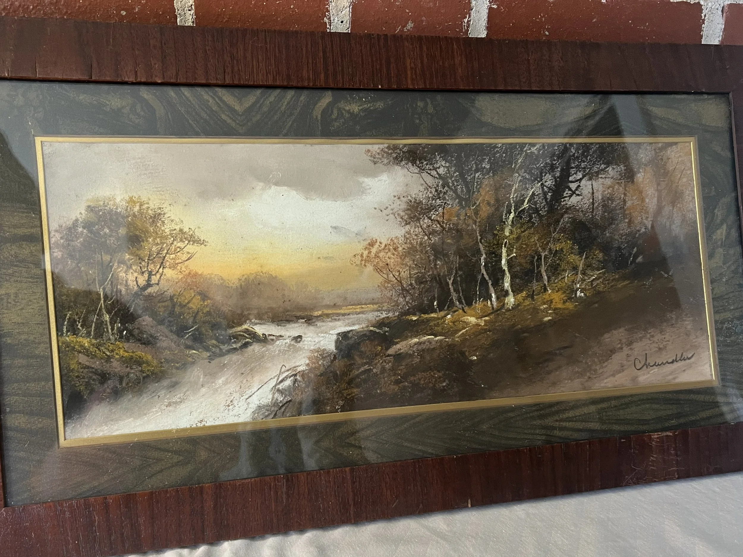 A framed landscape painting of a river flowing through trees with autumn foliage under a cloudy sky.