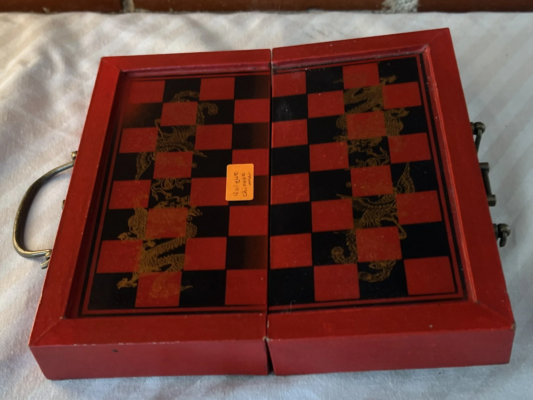 A vintage chess set with red and black checkered board decorated with gold dragons, housed in a red wooden case with a gold handle.