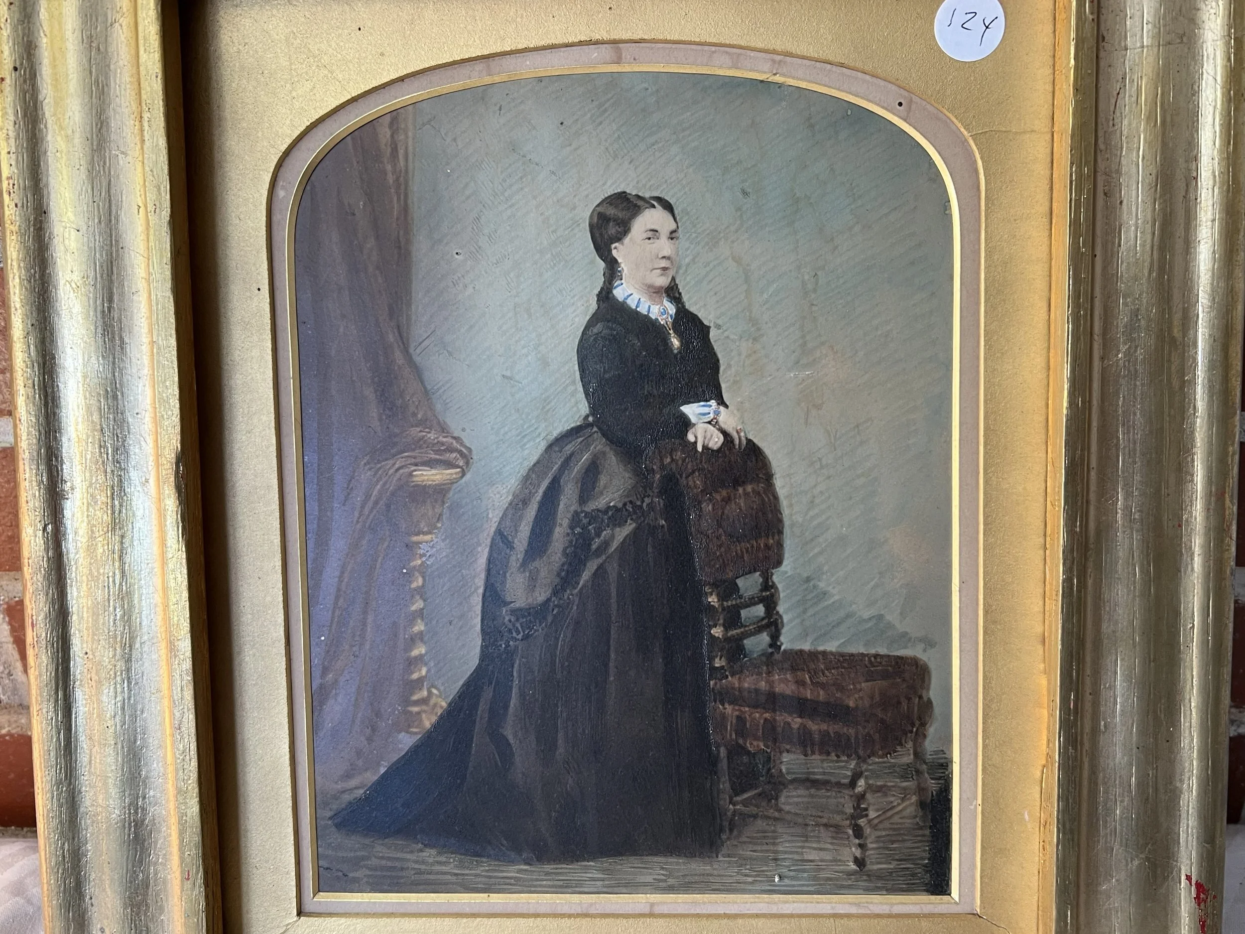 A portrait painting of a woman with dark hair, seated next to an ornate wooden chair, wearing a black dress with a blue and white collar, and accessorized with jewelry, framed with a gold border.