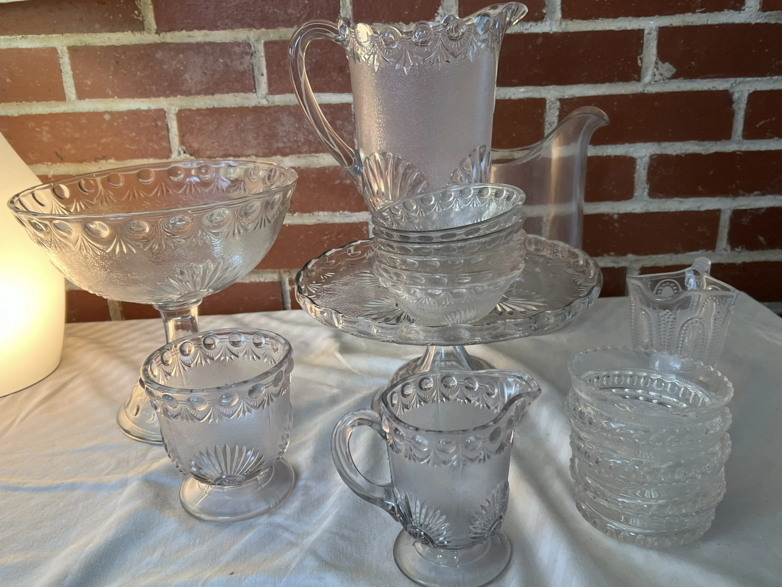 A collection of clear cut glass dishware, including bowls, cups, a large pitcher, a smaller pitcher, and a vase, arranged on a white tablecloth against a brick wall background.