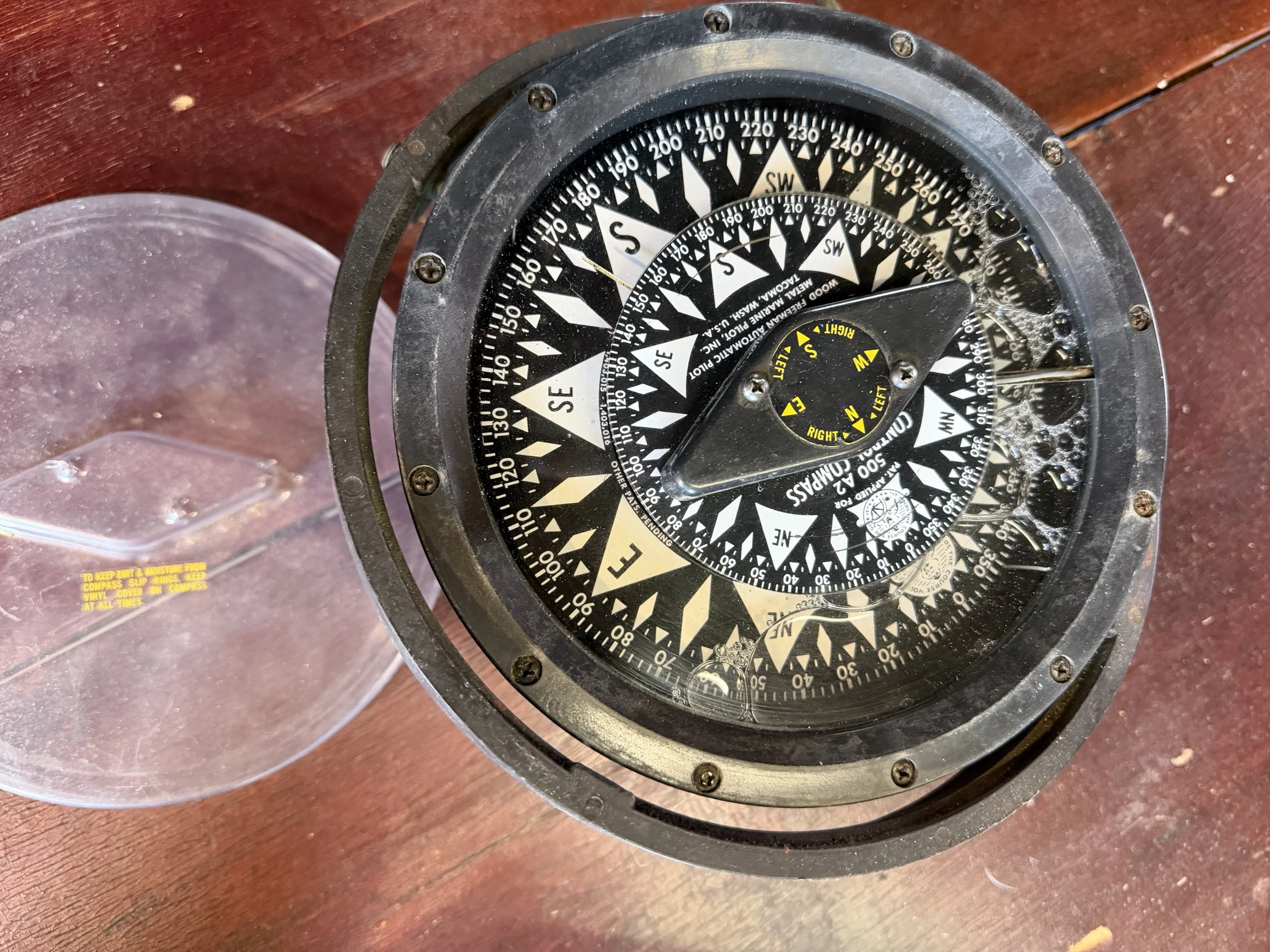A vintage aircraft compass mounted on a wooden surface, with a clear plastic cover to its side.