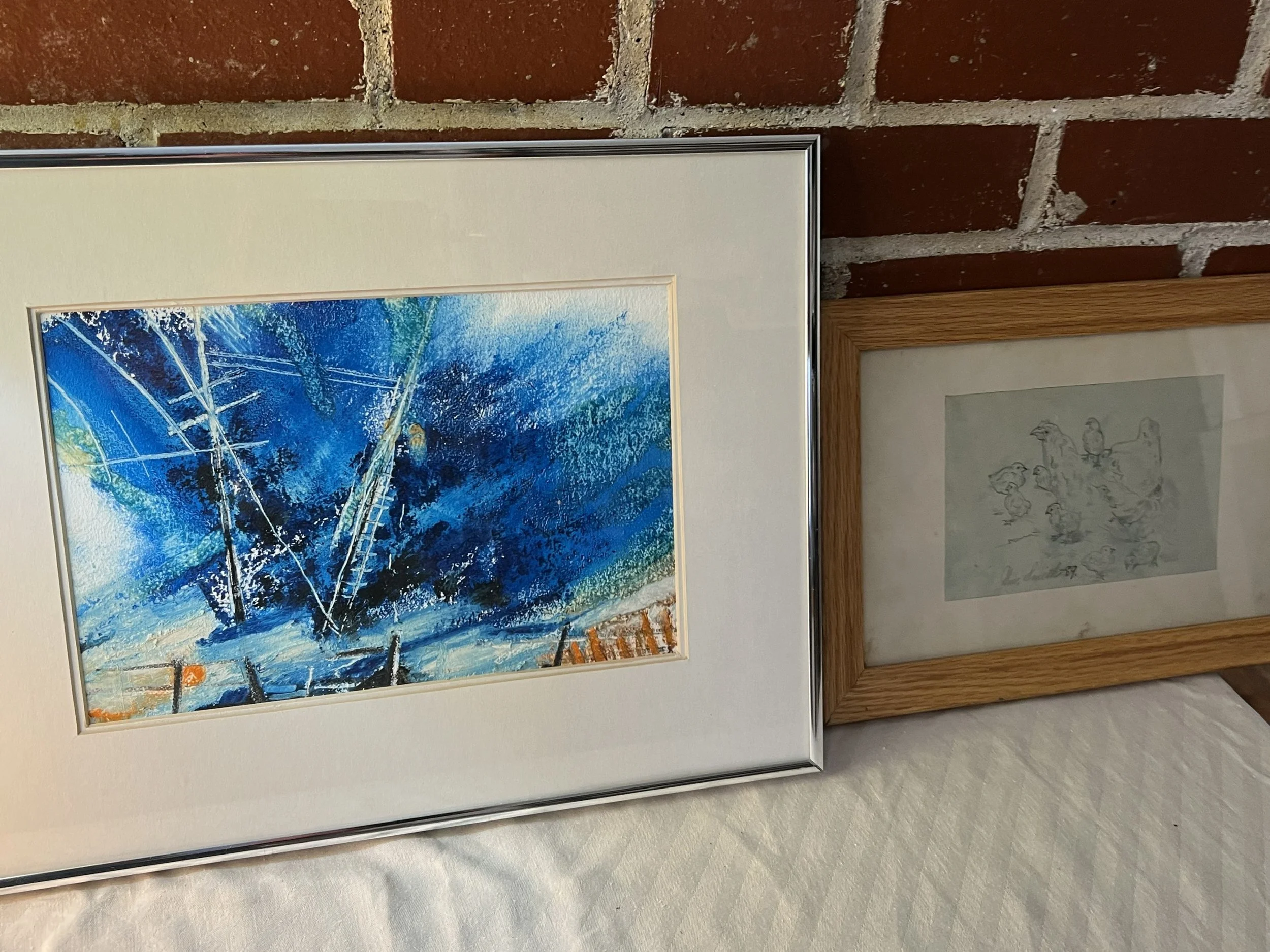 Two framed art pieces .The larger piece on the left is a colorful abstract painting with primarily blue tones, depicting a scene with trees and a snowy landscape. The smaller piece on the right is a black-and-white sketch or drawing of birds.