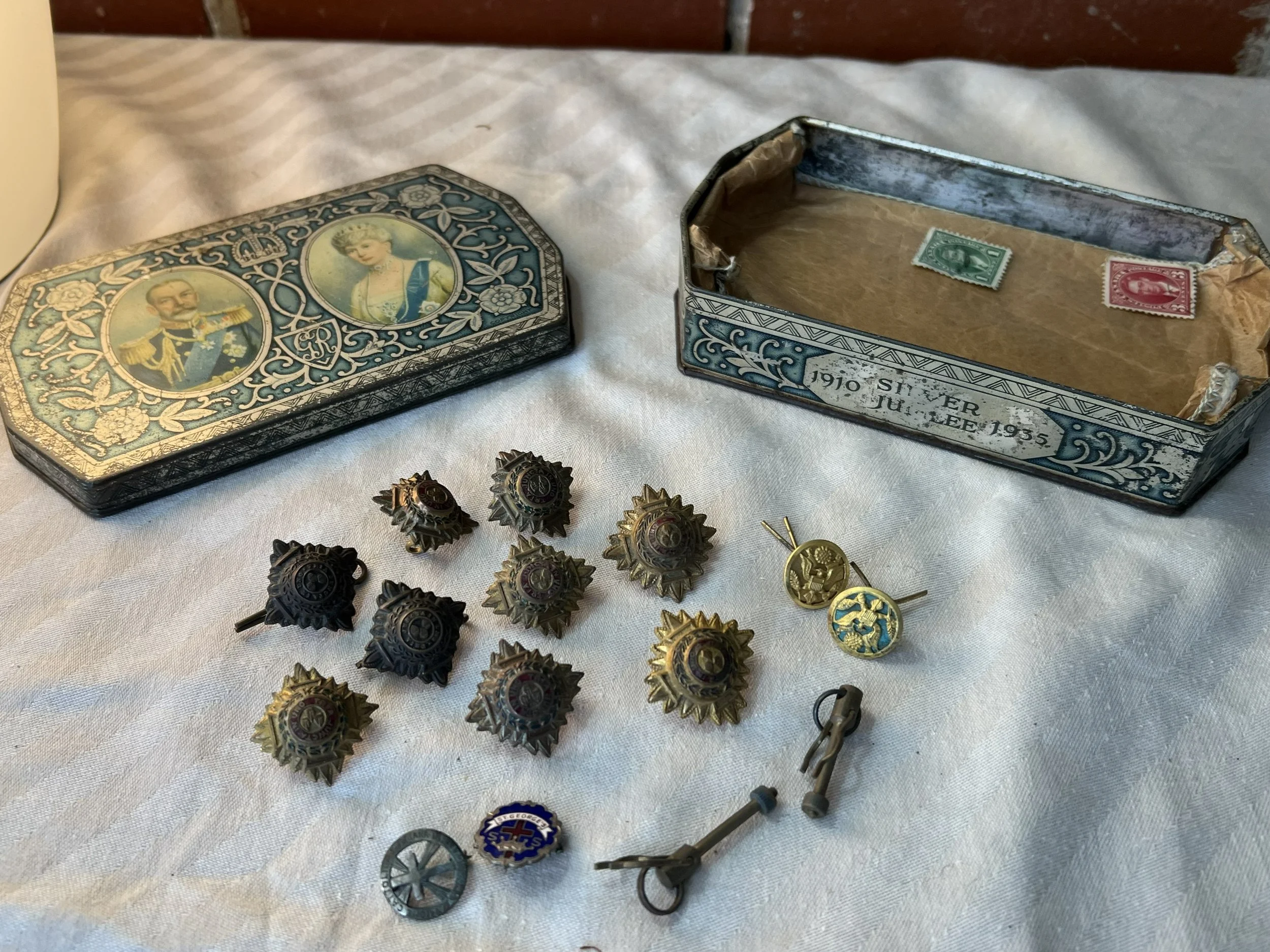 Vintage medals, pins, and badges on a white cloth, with an ornate metal tin box featuring portraits of a man and a woman and an empty decorative box with stamps inside.