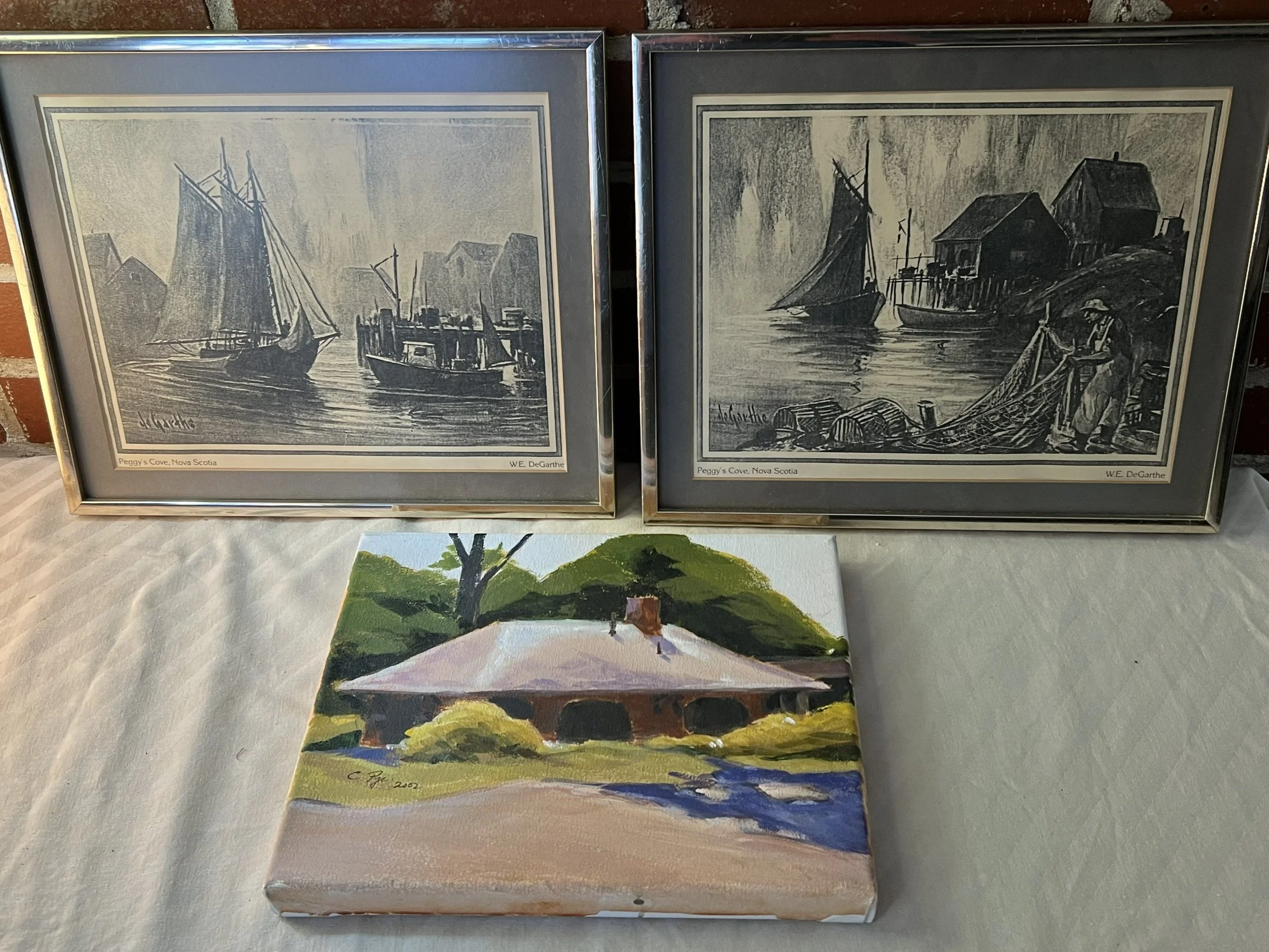 three collectible art pieces, including two framed prints and one canvas painting.
The two framed prints are artistic depictions of boats and fishing scenes at Peggy's Cove, Nova Scotia.
The small canvas painting Is the Annapolis Royal Train Station.