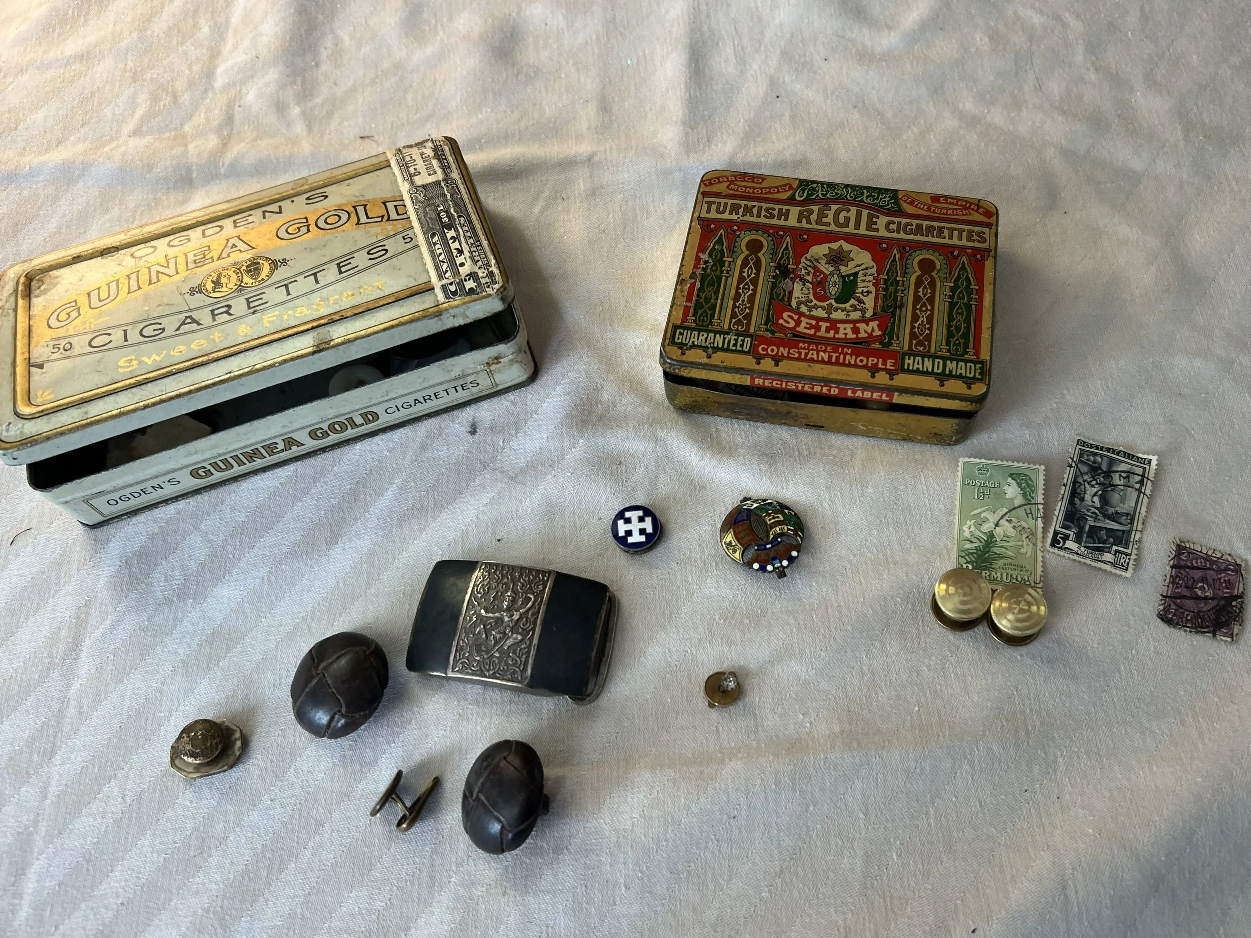 Vintage tobacco and cigarette tins, coins, jewelry, and small collectibles on a white cloth surface.