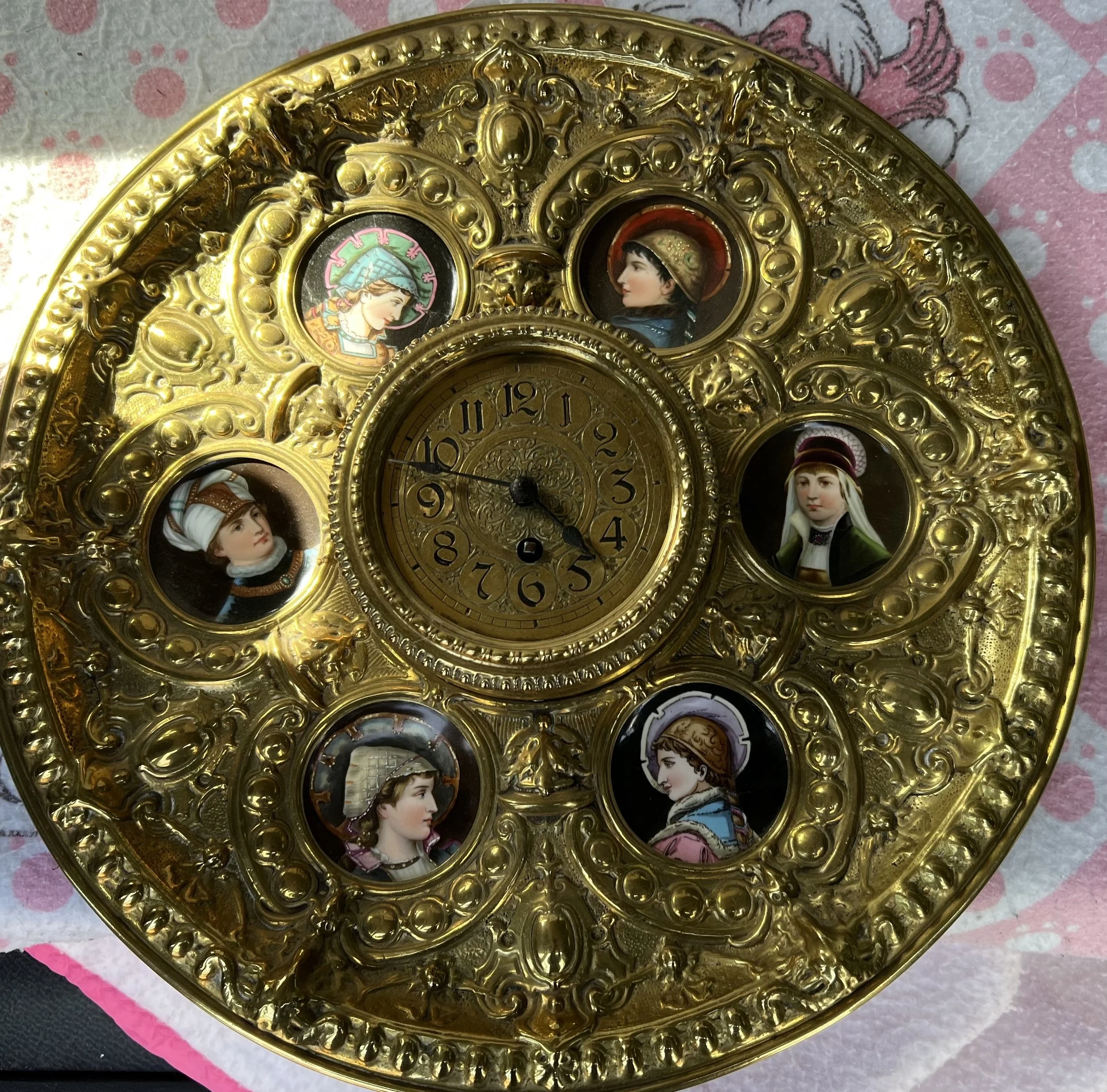 Ornate timepiece is a Victorian-style brass clock, often designed as a decorative mantle or desk piece.
The clock's most distinctive features are the six miniature portrait medallions surrounding the central dial. These portraits appear to be in the 
