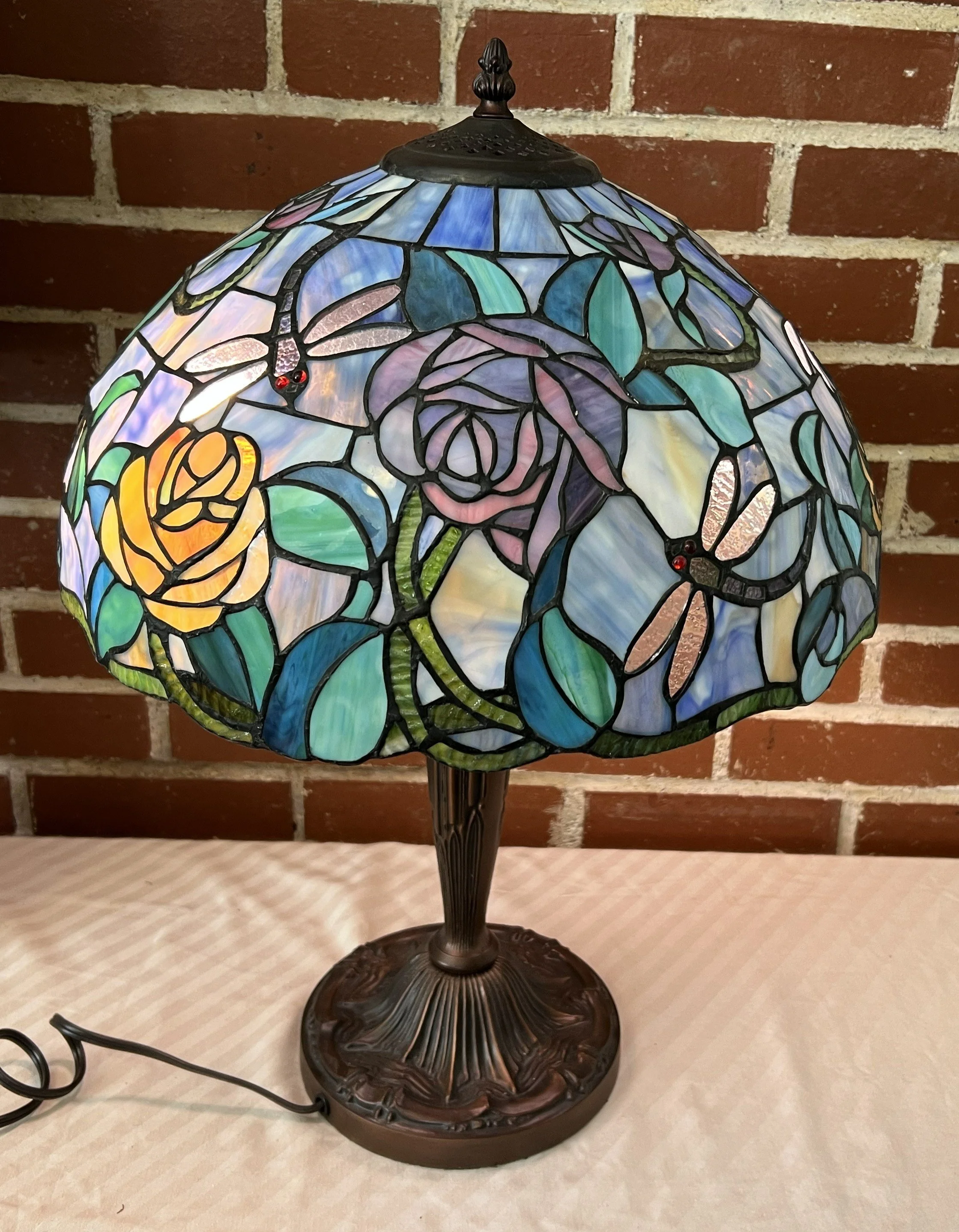 A stained glass Tiffany-style table lamp with a floral rose design, featuring purple, pink, yellow, blue, and green glass pieces, on a decorative bronze base against a brick wall background.