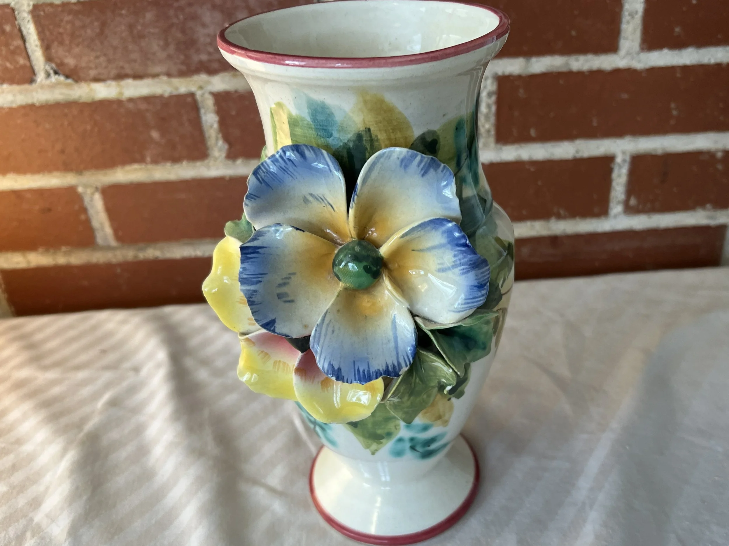 Colorful ceramic vase with a large yellow and blue flower decoration on the front, set against a brick wall background.