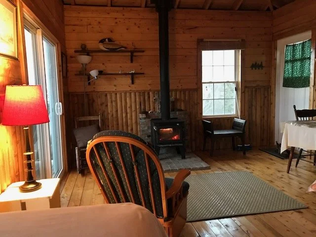 Cozy living room with wood-paneled walls, a wood-burning stove, a window, a sitting bench, a dining table, and a sliding glass door with a red lamp on a side table.