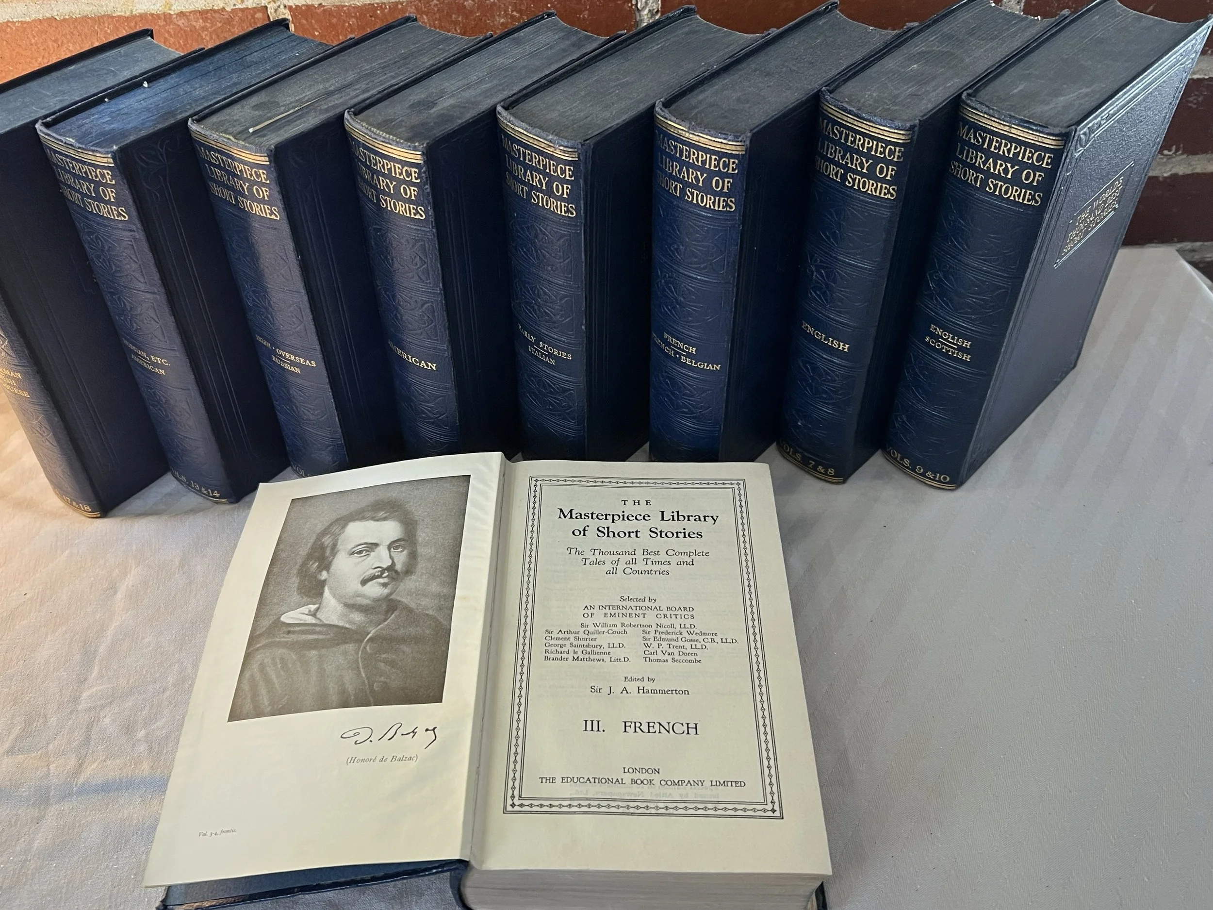 A row of hardcover books titled 'Masterpiece Library of Short Stories' standing upright on a table, with an open book displaying a portrait and the title page of 'The Masterpiece Library of Short Stories, III. French' in front.