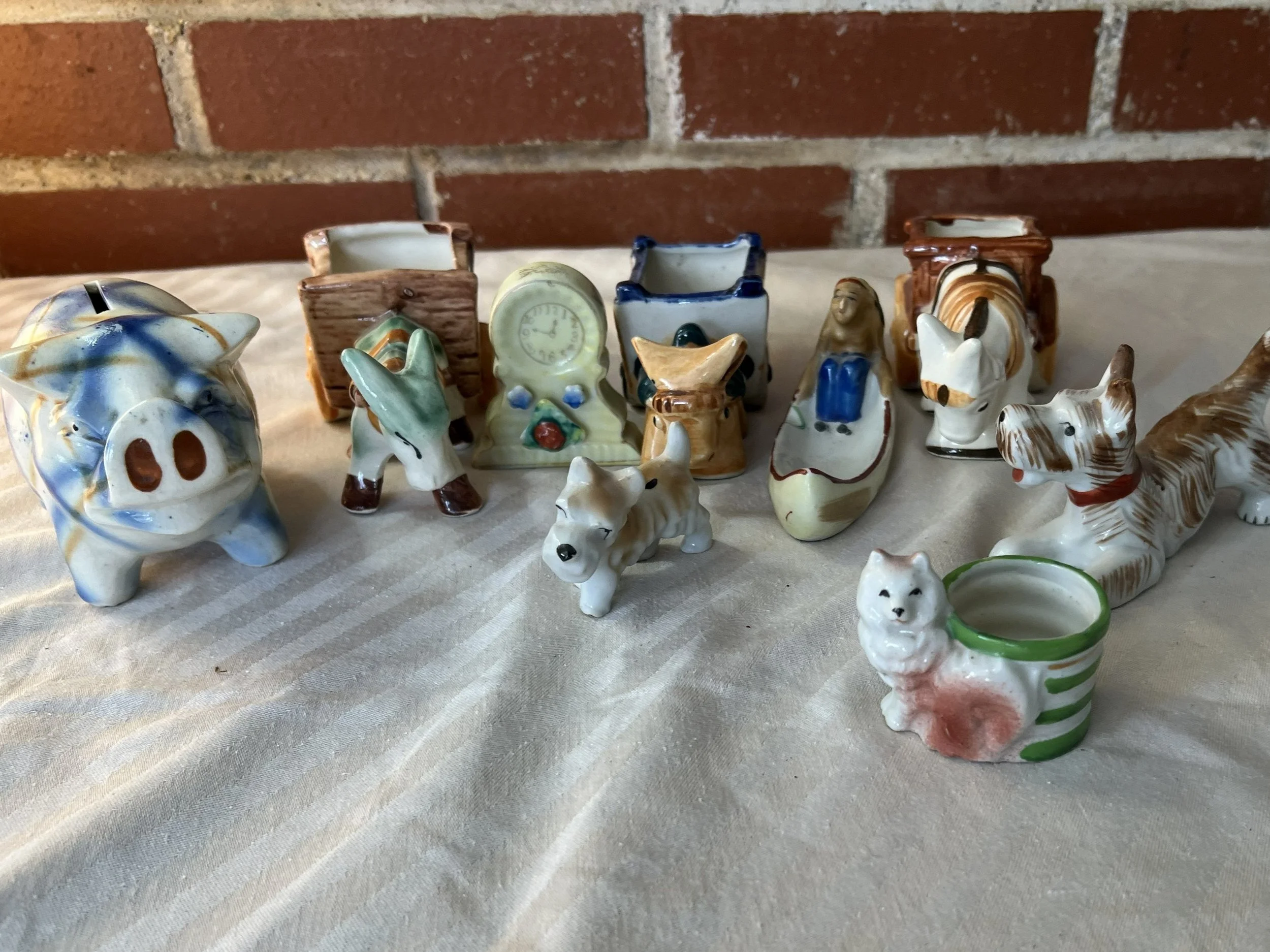 Collection of small ceramic animal figurines, including a pig, dogs, a horse, a cat, and various decorative containers, arranged on a light-colored fabric surface against a brick wall background.