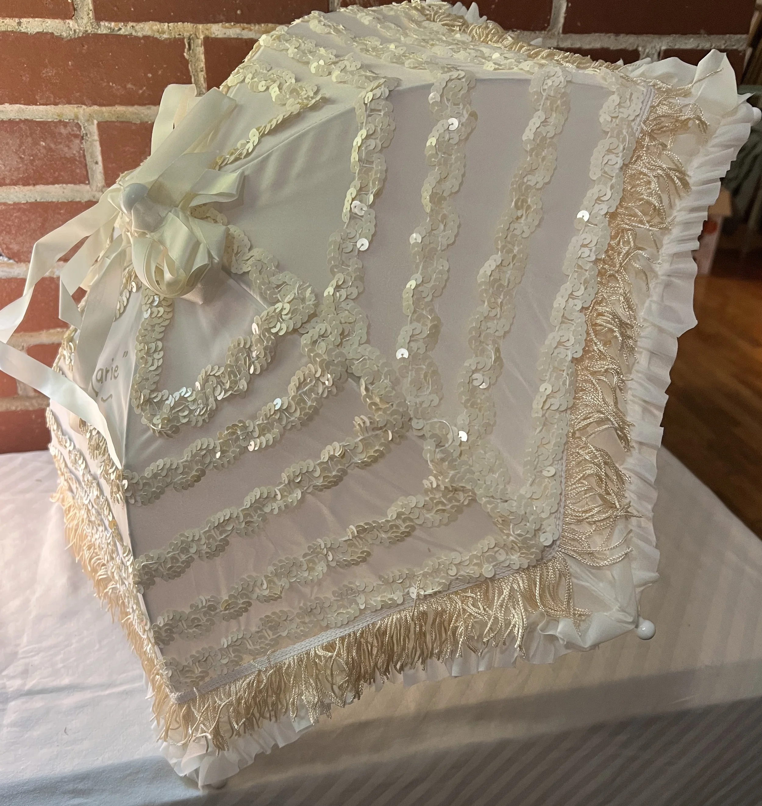 A white wedding dress box decorated with gold sequins, ribbons, and fringe, placed on a table against a brick wall.