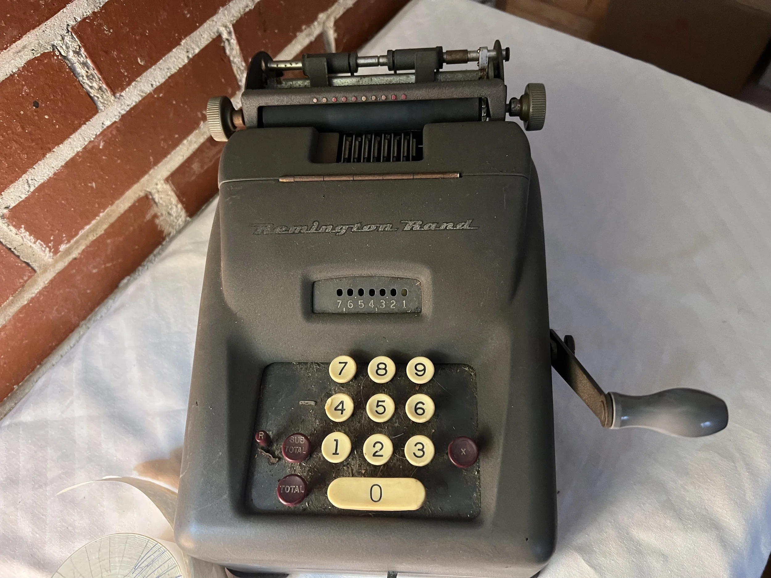 A vintage Remington Rand mechanical adding and calculating machine, likely dating from the 1940s or 1950s. 
Type: 10-key manual mechanical calculator. 
Features: Includes a printing mechanism for paper tape, a digit indicator window, and a hand crank